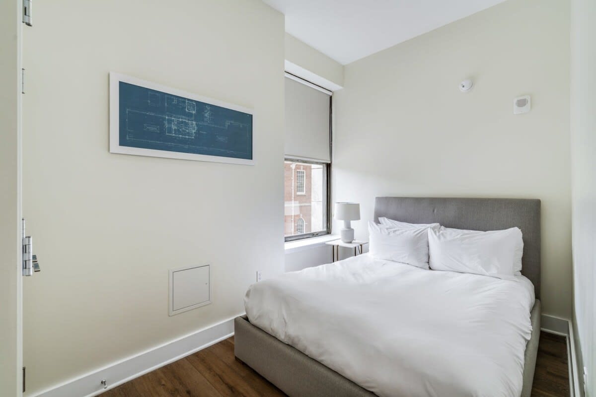 Cozy bedroom featuring a gray upholstered queen-size bed with white bedding, a small white nightstand with a lamp, and a window with a roller shade.