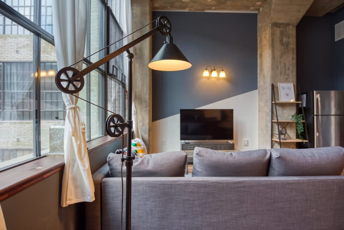 A close-up view of the grey sectional sofa in the living room, highlighting a unique industrial-style floor lamp with a pulley system, positioned next to large windows with white curtains.