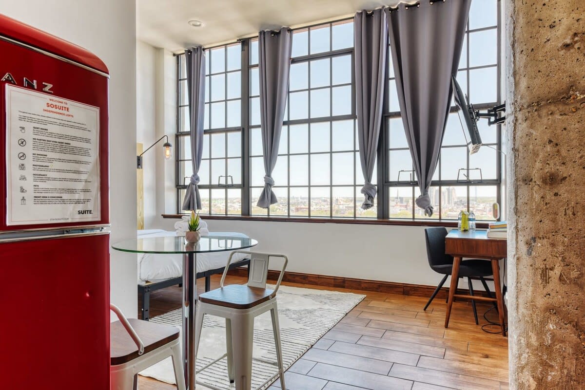 Wide view of the studio apartment showcasing a queen bed, a glass-top dining table with two stools, and a desk area by the large industrial windows. A vibrant red retro refrigerator adds a pop of color to the space.