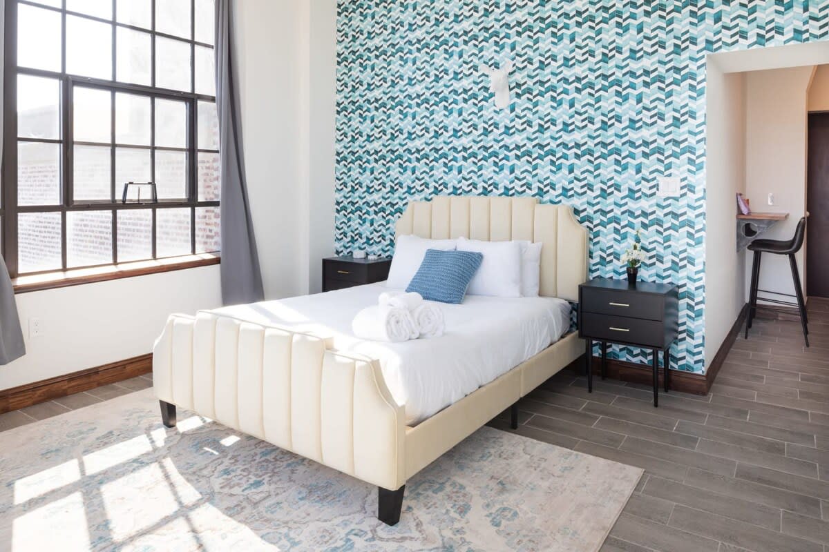 Cozy studio bedroom with a cream-colored upholstered bed set against a vibrant blue and white geometric patterned accent wall, flanked by black nightstands. A large window with dark frames and grey curtains is visible on the left.