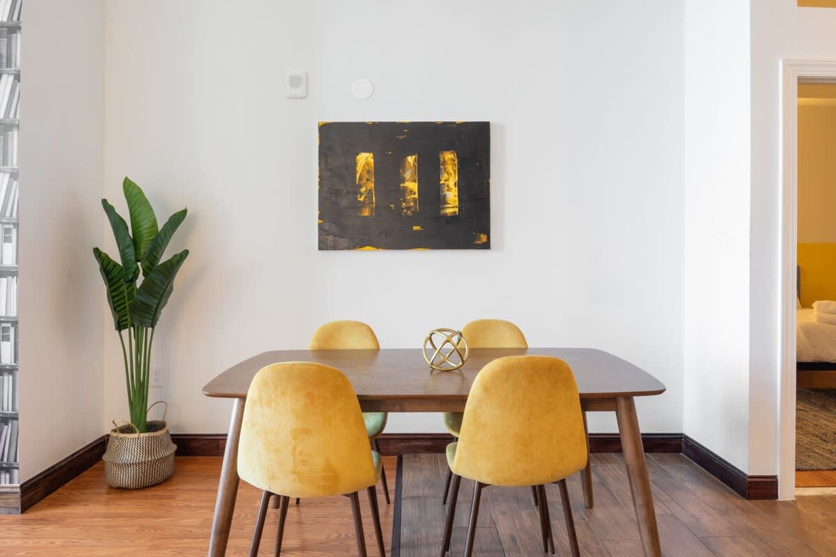 Dining area with a rectangular wooden table and four yellow velvet chairs, accented by a modern abstract painting on the wall and a potted plant in the corner.