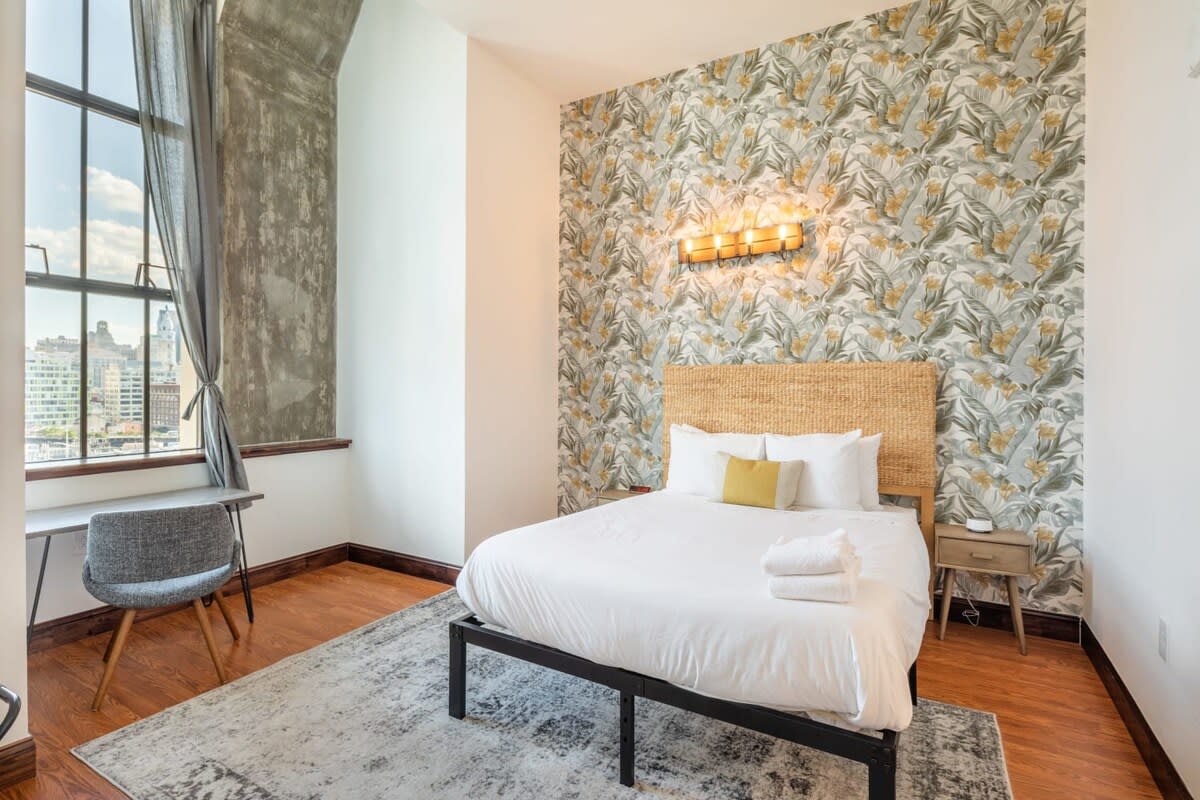 Bright bedroom featuring a queen-sized bed with a woven headboard, a small nightstand, and a desk with a chair, set against a floral wallpaper accent wall and large industrial windows.