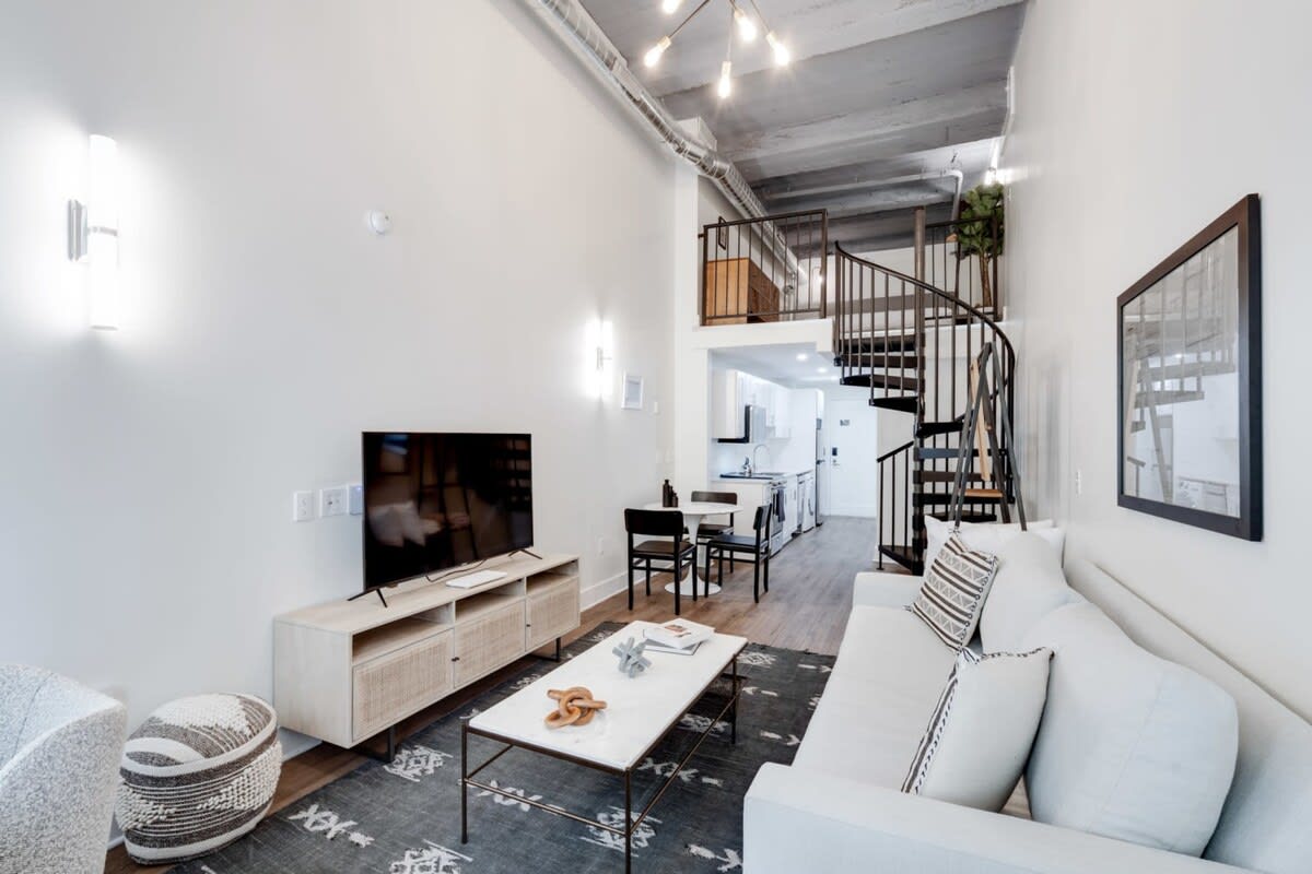 A spacious, open-concept living room with a light grey sofa, marble-top coffee table, and TV console, featuring a striking black spiral staircase leading to an upper loft and an adjacent dining area and kitchen.