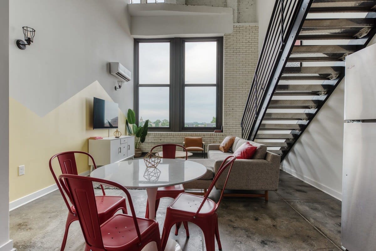 Bright living and dining area with a white round dining table and red metal chairs, a light gray sectional sofa, and large windows offering an expansive view. A white console table with a TV is positioned against a wall with a yellow geometric accent.