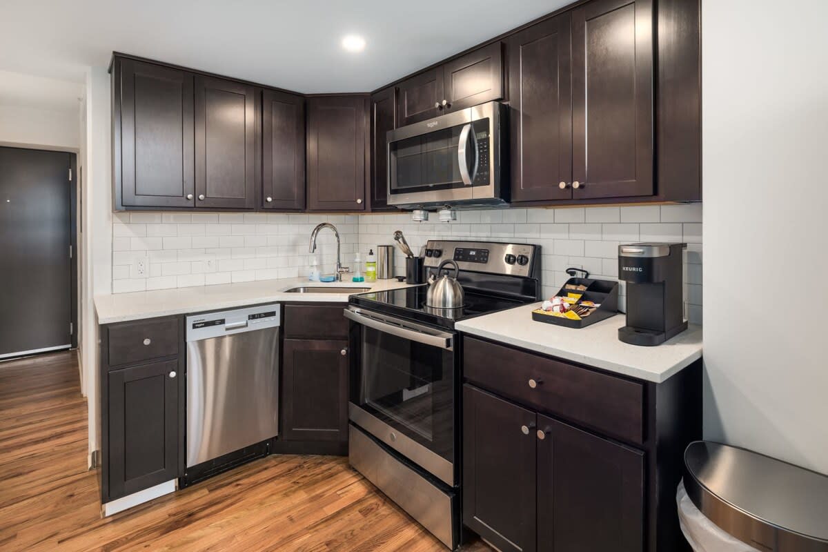 A well-equipped kitchen with dark wood cabinetry, white subway tile backsplash, and light countertops. Stainless steel appliances include a microwave, stove, dishwasher, and a Keurig coffee maker on the counter.