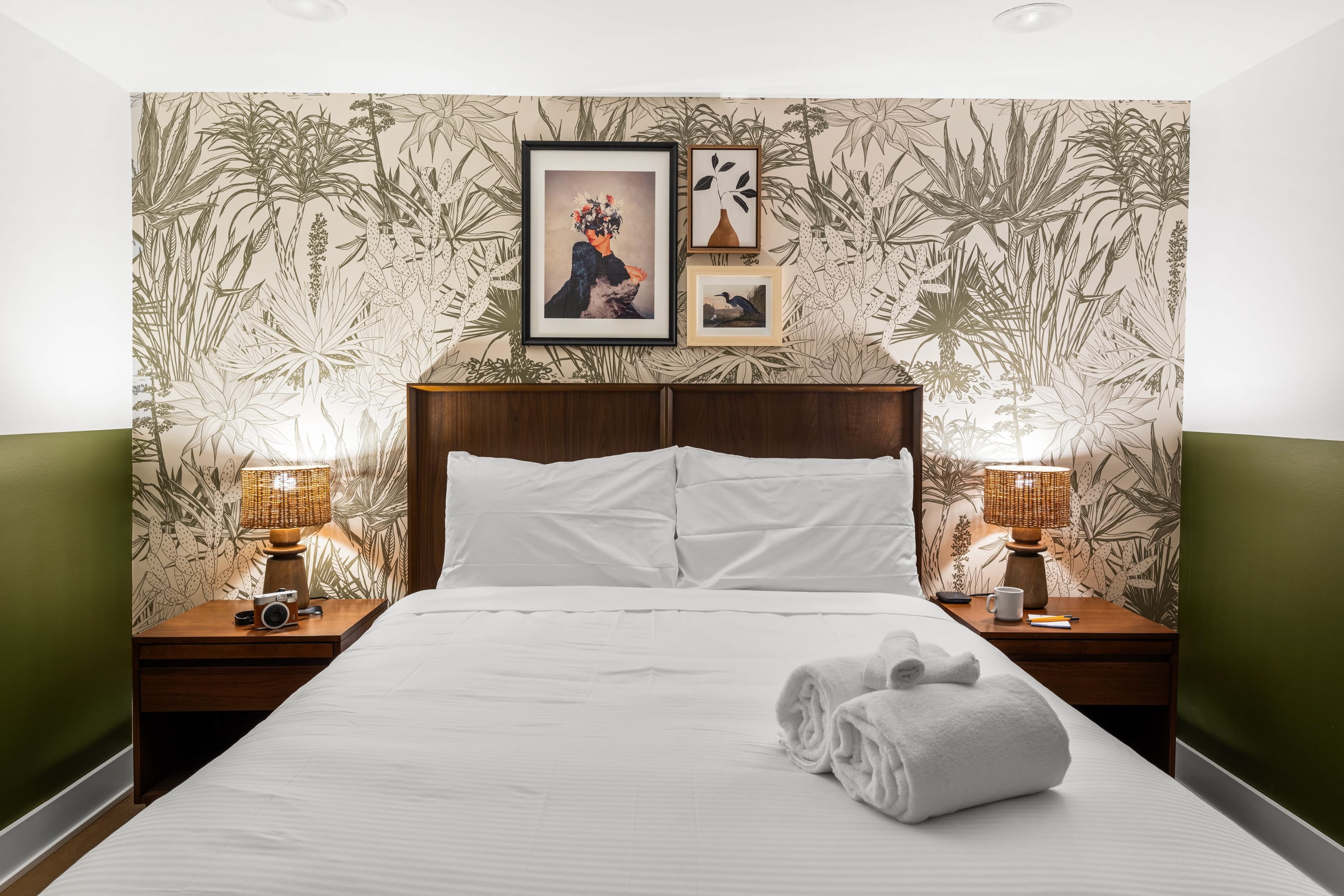 A cozy bedroom with a queen-sized bed featuring white linens and a dark wood headboard, flanked by two wooden nightstands with woven lamps. The accent wall behind the bed has a botanical wallpaper, and the side walls are olive green.