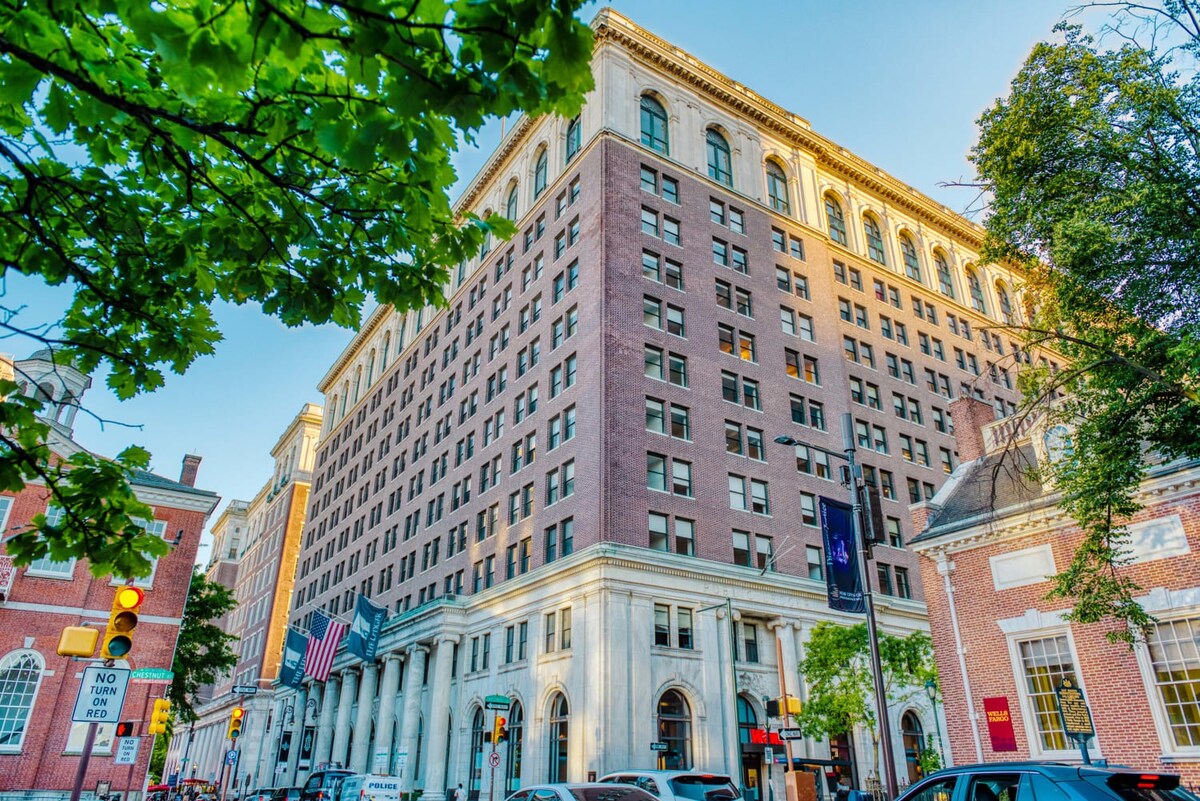 A grand, multi-story brick building with classical architectural details, including columns and arched windows, surrounded by green trees under a clear sky.