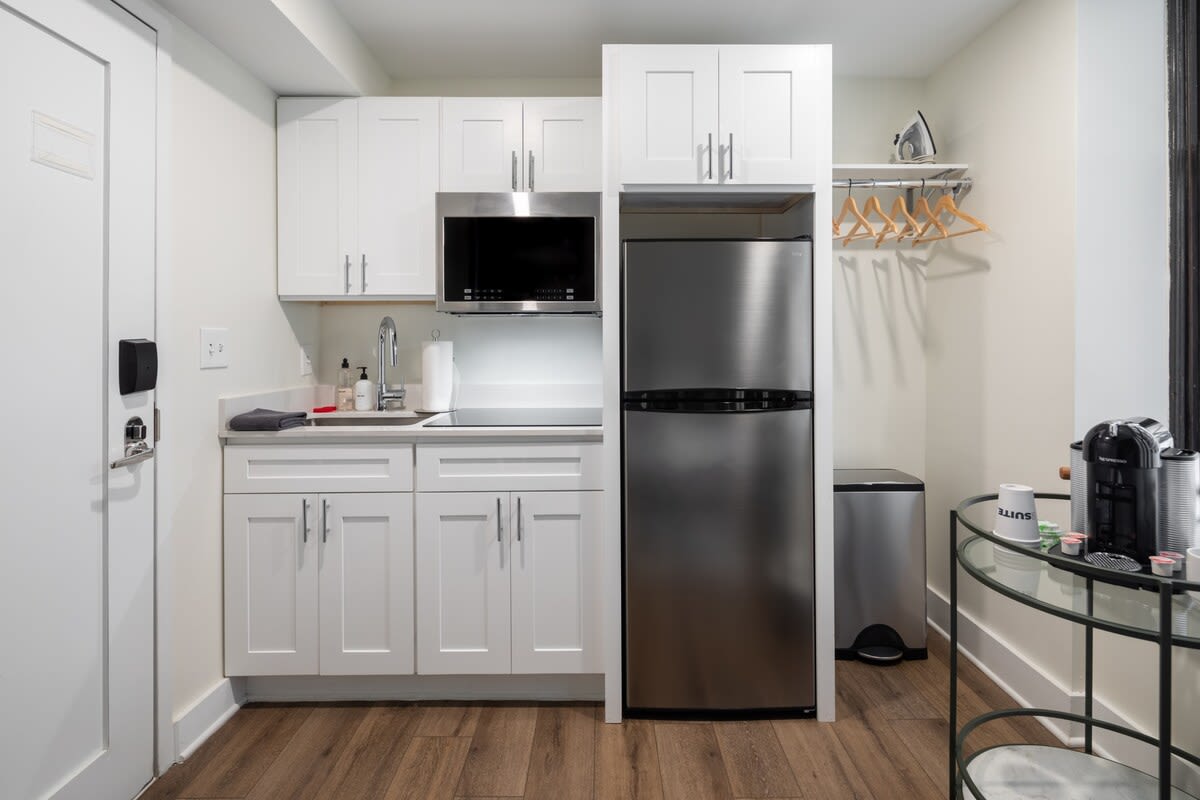 A compact, modern kitchenette featuring white shaker cabinets, stainless steel appliances including a microwave and a two-door refrigerator, and a small sink with a two-burner cooktop. A stainless steel trash can and a hanging rack with hangers are visible next to the fridge.