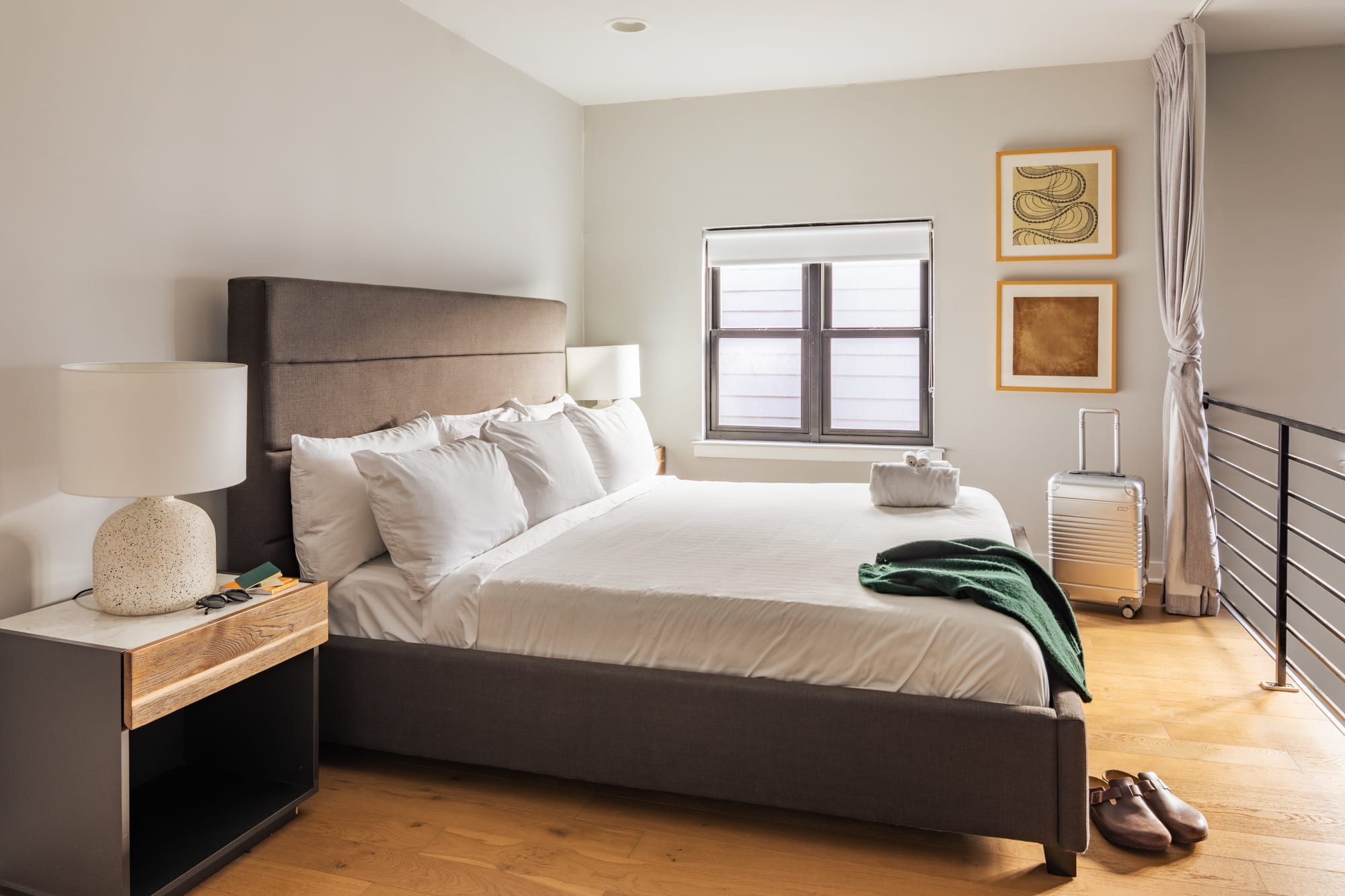 A bright bedroom with a large bed featuring a dark gray upholstered headboard, white bedding, and a wooden nightstand with a white lamp. Two framed abstract artworks hang above the window, and a metal railing suggests a loft space.