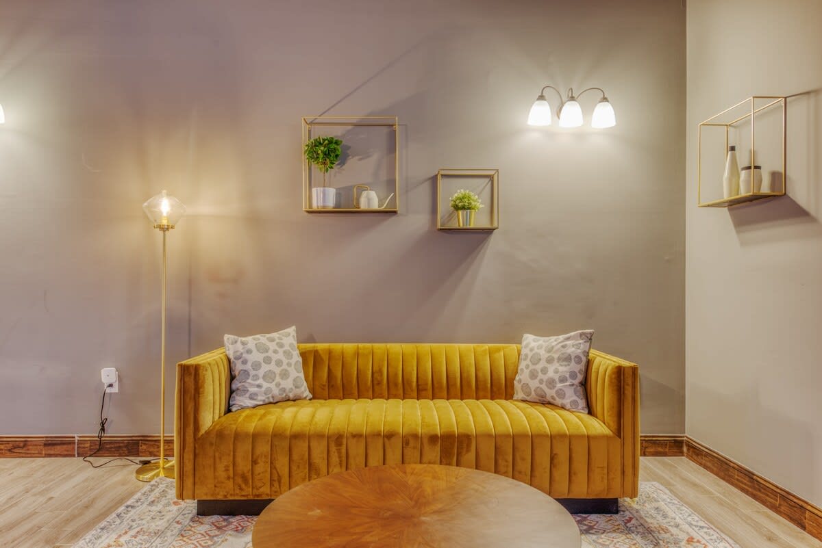 A plush mustard yellow channel-tufted sofa with patterned throw pillows is centered on an ornate rug, flanked by a modern floor lamp and wall-mounted gold cube shelves with greenery.