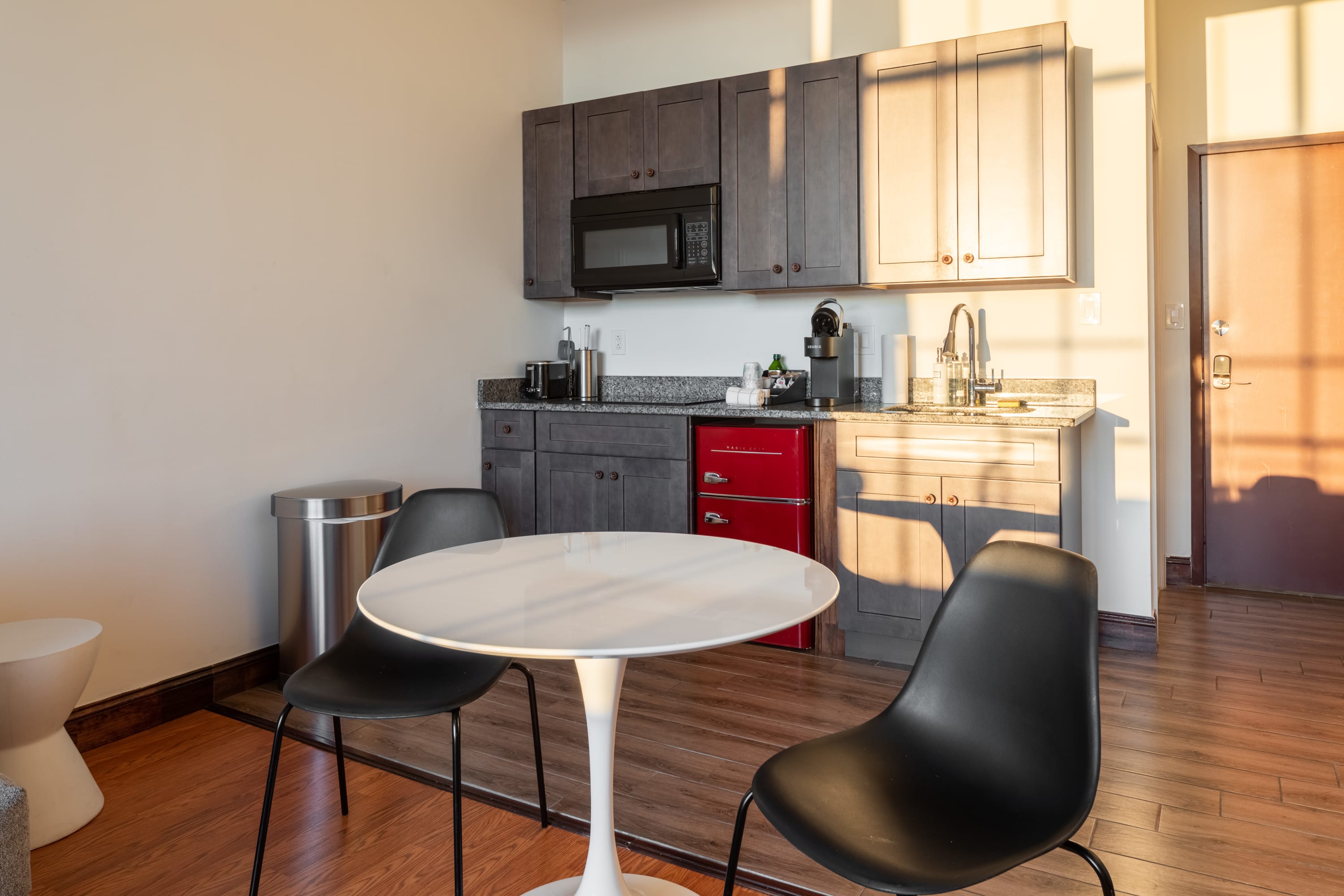 A compact kitchen area featuring dark gray shaker cabinets, granite countertops, a black microwave, and a vibrant red mini-fridge, paired with a small white round dining table and two black modern chairs.