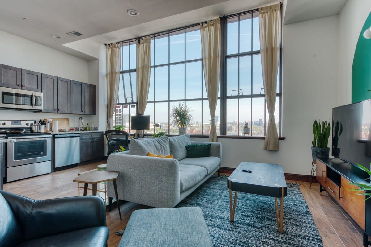 An open-concept living room and kitchen area features a light gray sofa, a black coffee table, and a kitchen with dark gray cabinets and stainless steel appliances. Large industrial windows with city views dominate the space, and a black leather armchair is visible in the foreground.