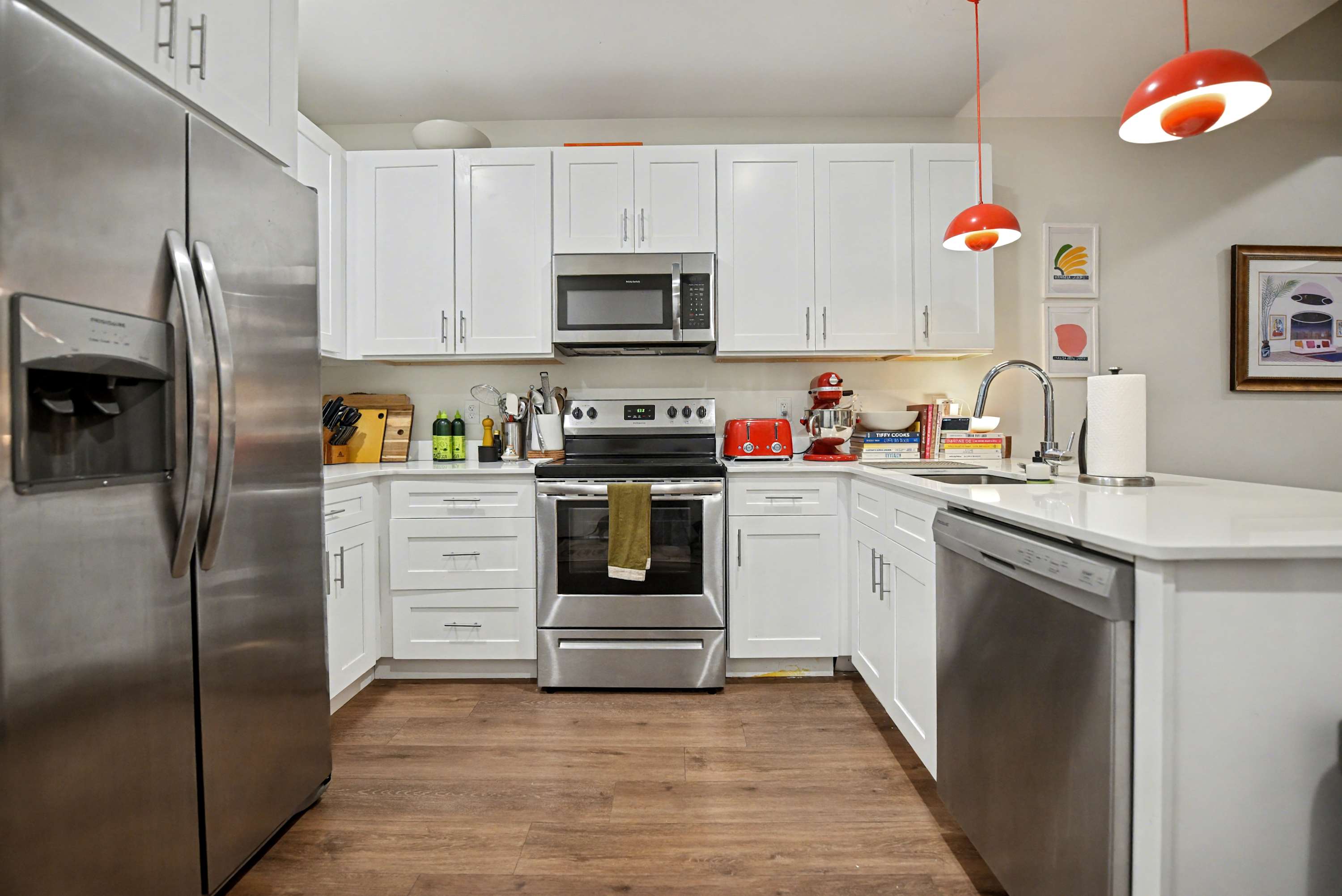 A bright, modern kitchen features white shaker cabinets, white countertops, and stainless steel appliances including a refrigerator, oven, microwave, and dishwasher. Red kitchen appliances add pops of color.