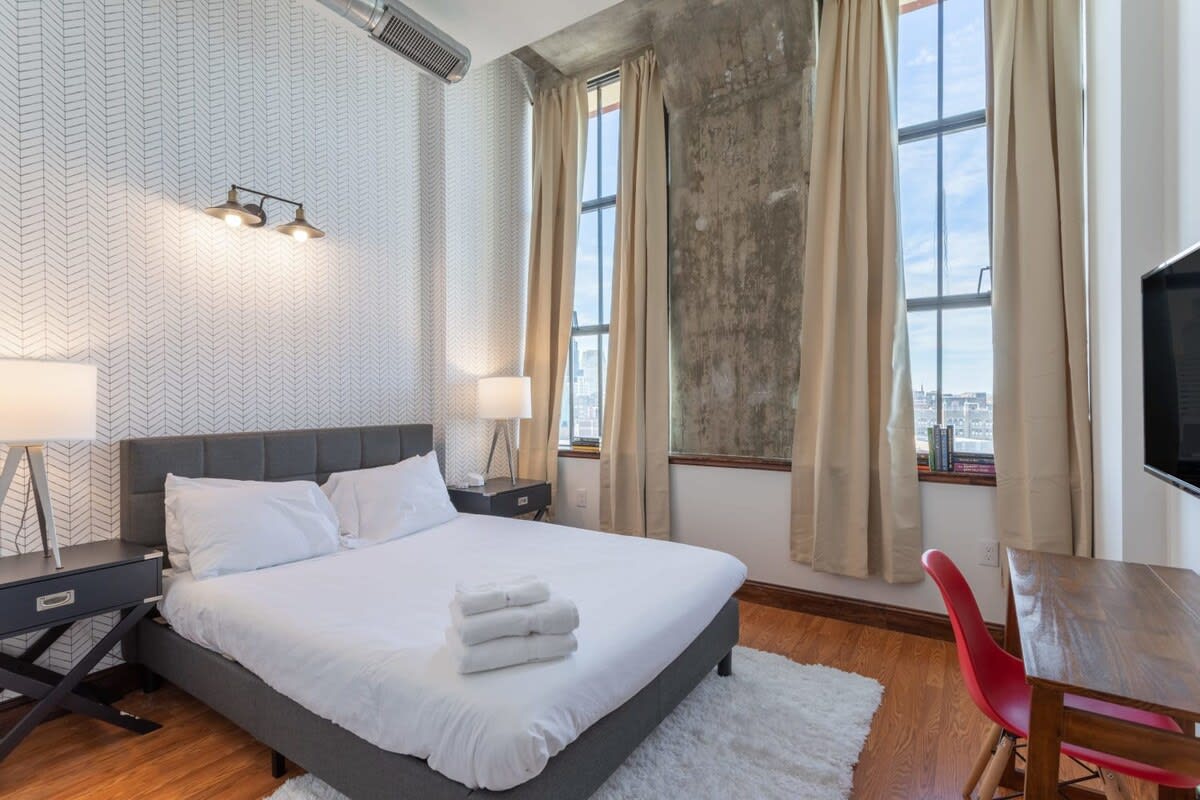 A cozy bedroom featuring a queen-sized bed with a dark gray upholstered headboard and a subtle herringbone patterned wallpaper. Large industrial windows with beige curtains overlook a city landscape.