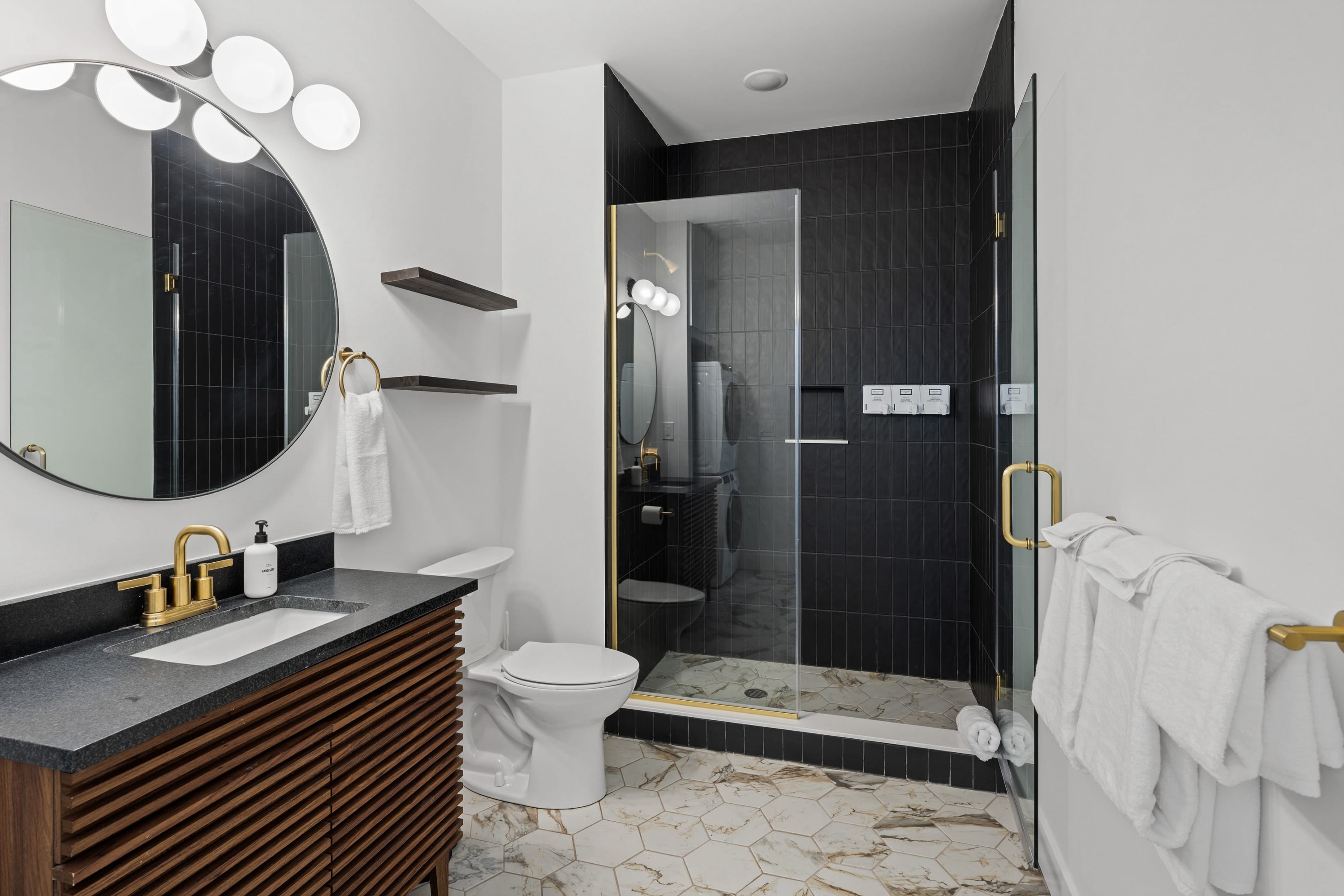 Stylish bathroom featuring a dark vanity with gold fixtures, a round mirror with circular lights, and a walk-in shower with black subway tiles and a clear glass enclosure.