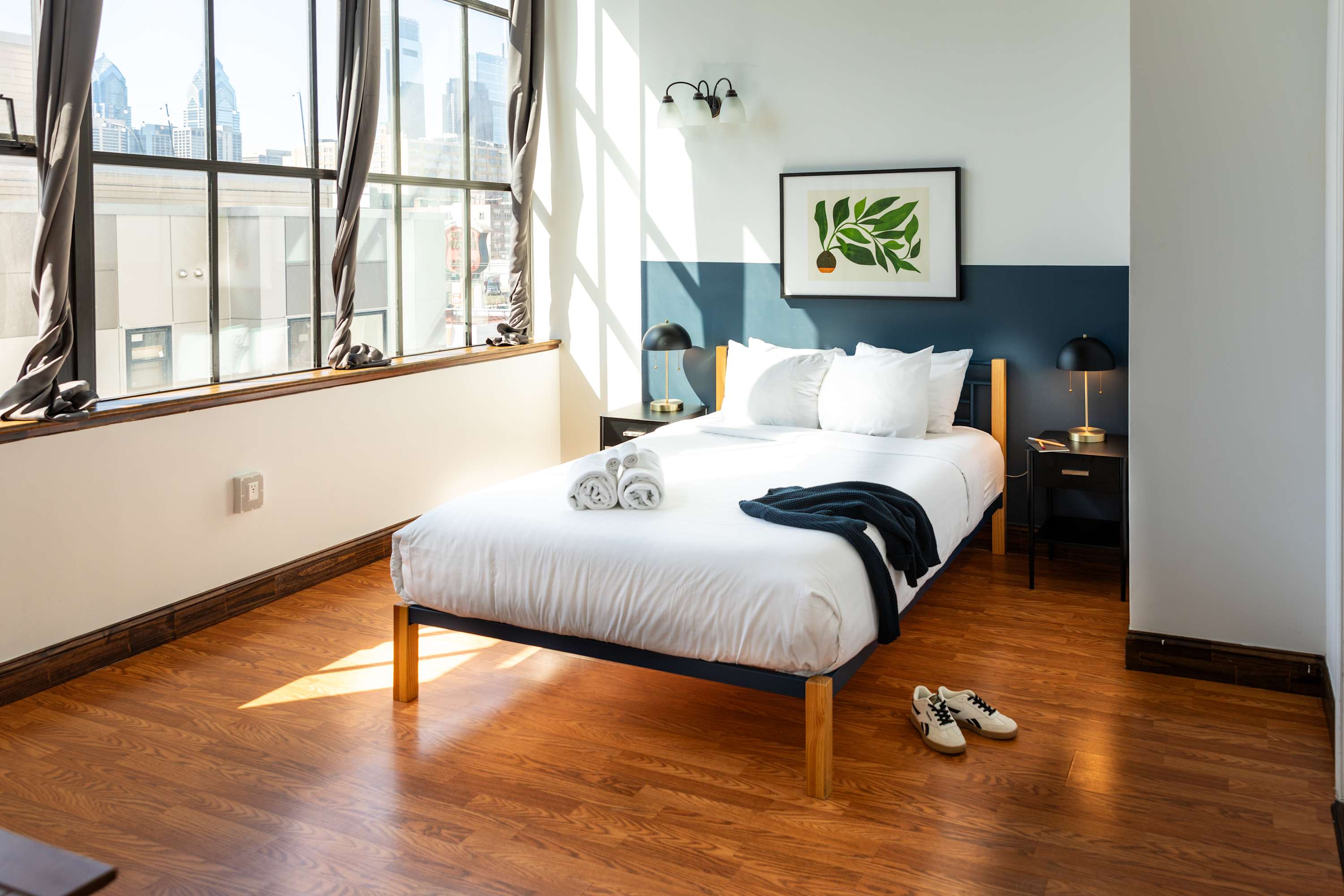 Sunlit bedroom with a white bed featuring a light wood and black frame, flanked by black nightstands and lamps. Large industrial-style windows offer city views, and the wall has a white and dark blue two-tone design.