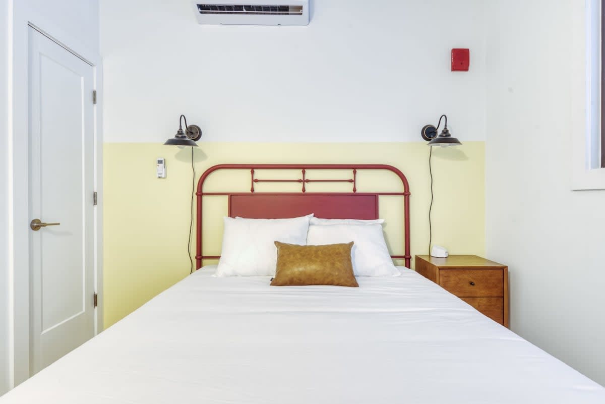 Cozy bedroom with a red metal-frame bed, white bedding, and a brown accent pillow, flanked by wall-mounted sconces and a wooden nightstand, set against a two-toned white and yellow wall.