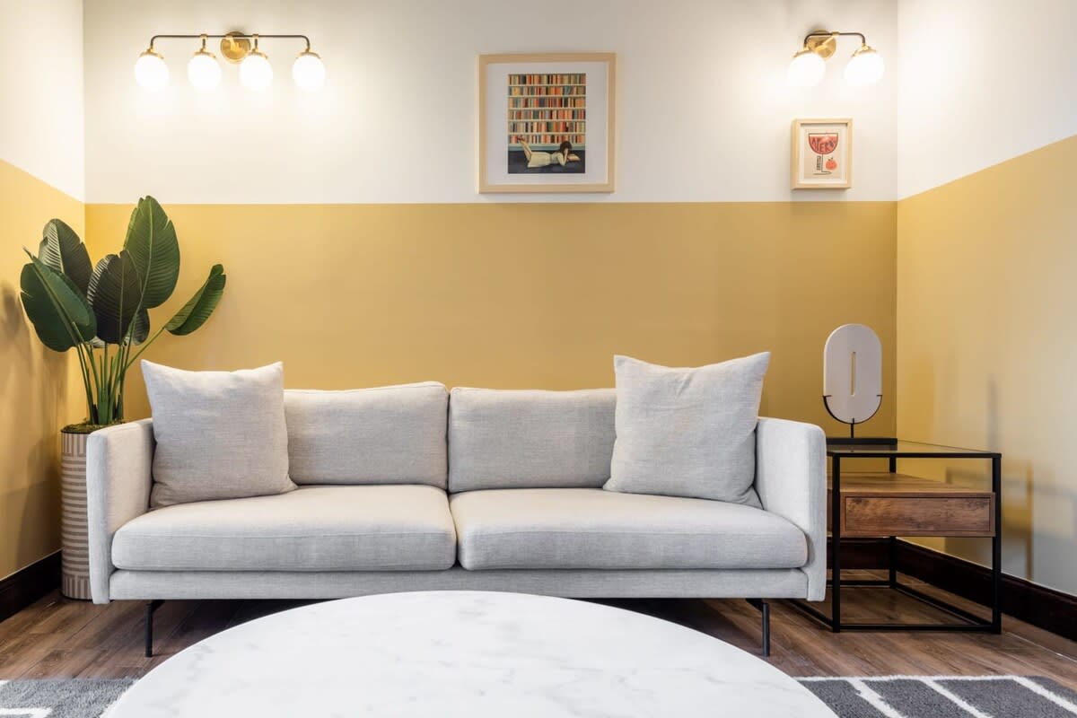 A cozy living room corner featuring a light gray sofa with throw pillows against a two-tone wall painted white and mustard yellow. Modern wall sconces and framed artwork adorn the wall above the sofa.