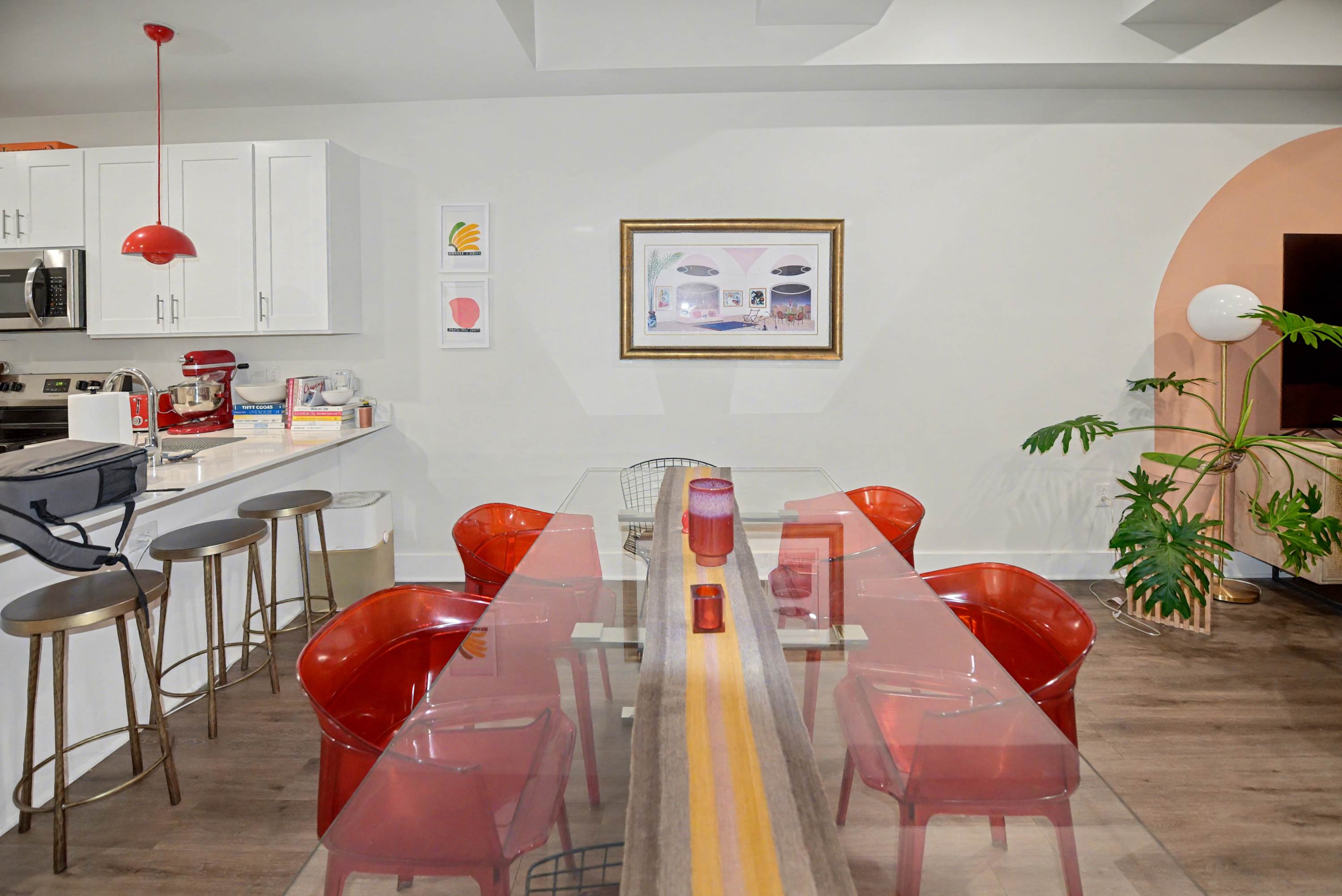 A modern dining area with a glass-top table and red transparent chairs is positioned between a white kitchen with stainless steel appliances and a living area with a peach arch accent wall. Red pendant lights hang above the kitchen island.