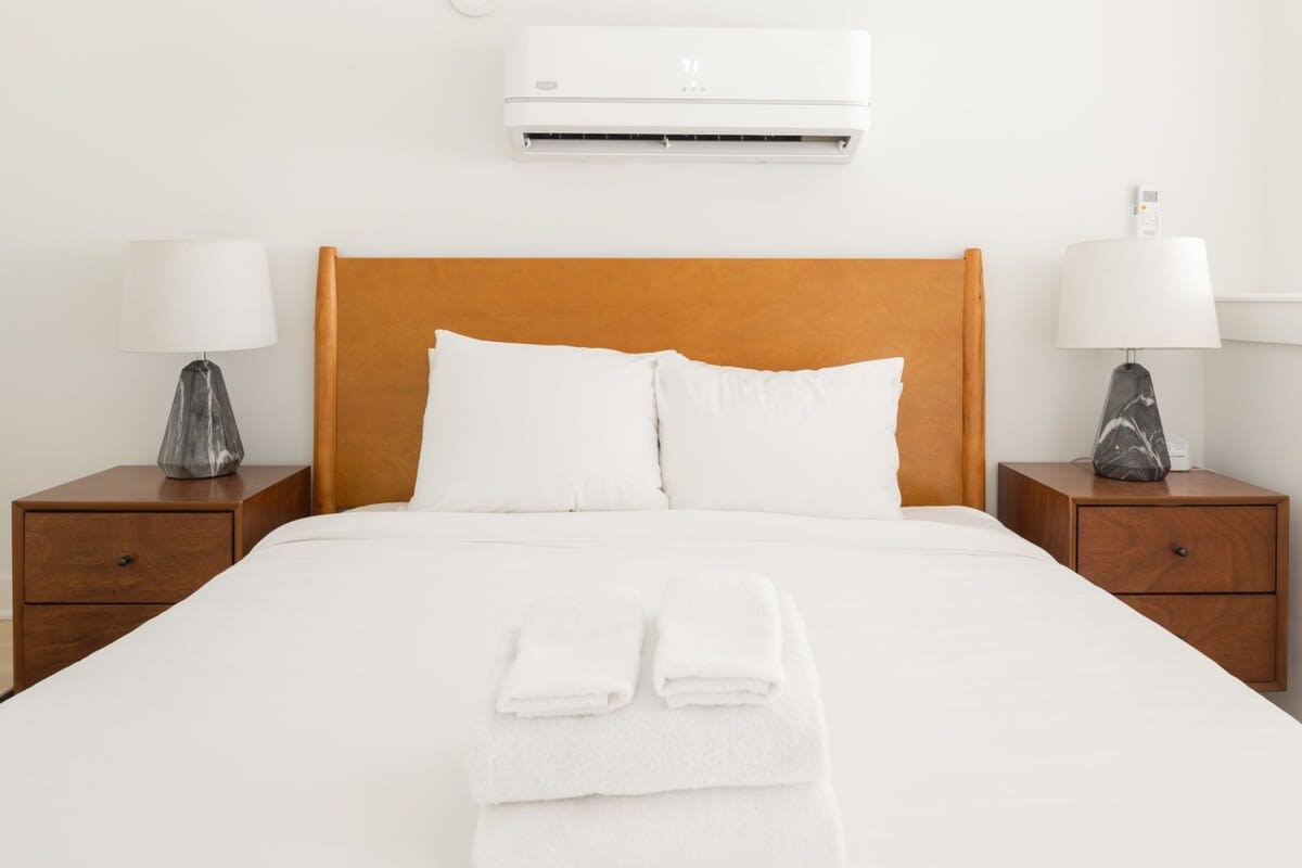 A cozy bedroom with a queen-sized bed featuring a light brown leather headboard, white bedding, and two white pillows. Two dark wood nightstands flank the bed, each topped with a gray and white patterned lamp. An air conditioning unit is mounted above the bed.