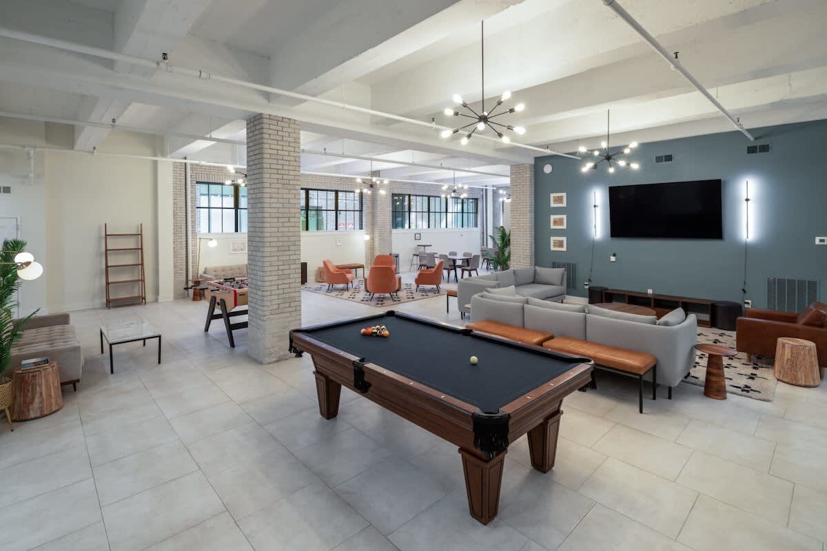 A spacious communal lounge area with multiple seating arrangements, including sofas and armchairs, and a large pool table in the foreground. The room features exposed brick columns, large windows, and modern chandeliers.