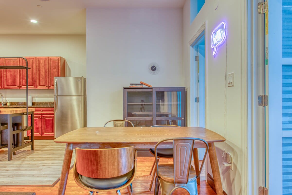 Dining area with a light wood table and four chairs, including two with wooden backs and two with metal frames. A black storage cabinet and a neon 'hello' sign are in the background.
