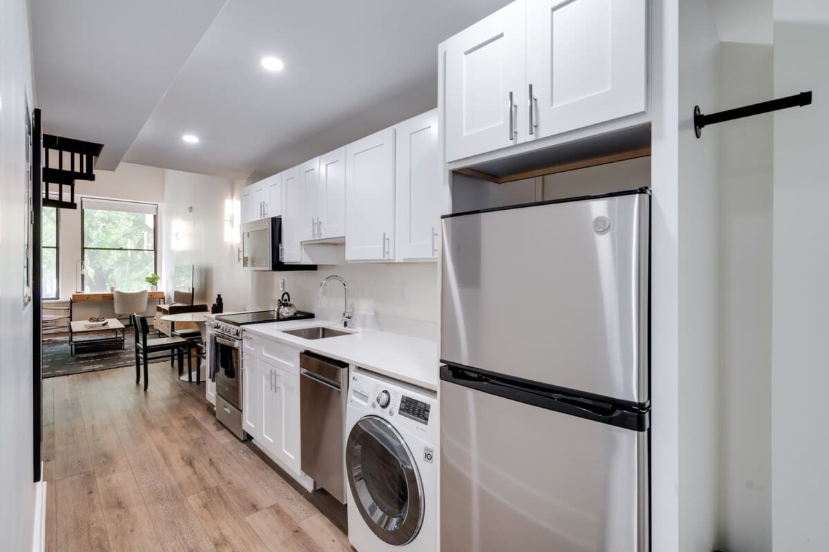 Compact, modern galley kitchen with white shaker-style cabinets, stainless steel appliances including a refrigerator, stove, dishwasher, and a washer/dryer combo. White countertops and light wood-look flooring extend into the open living area.