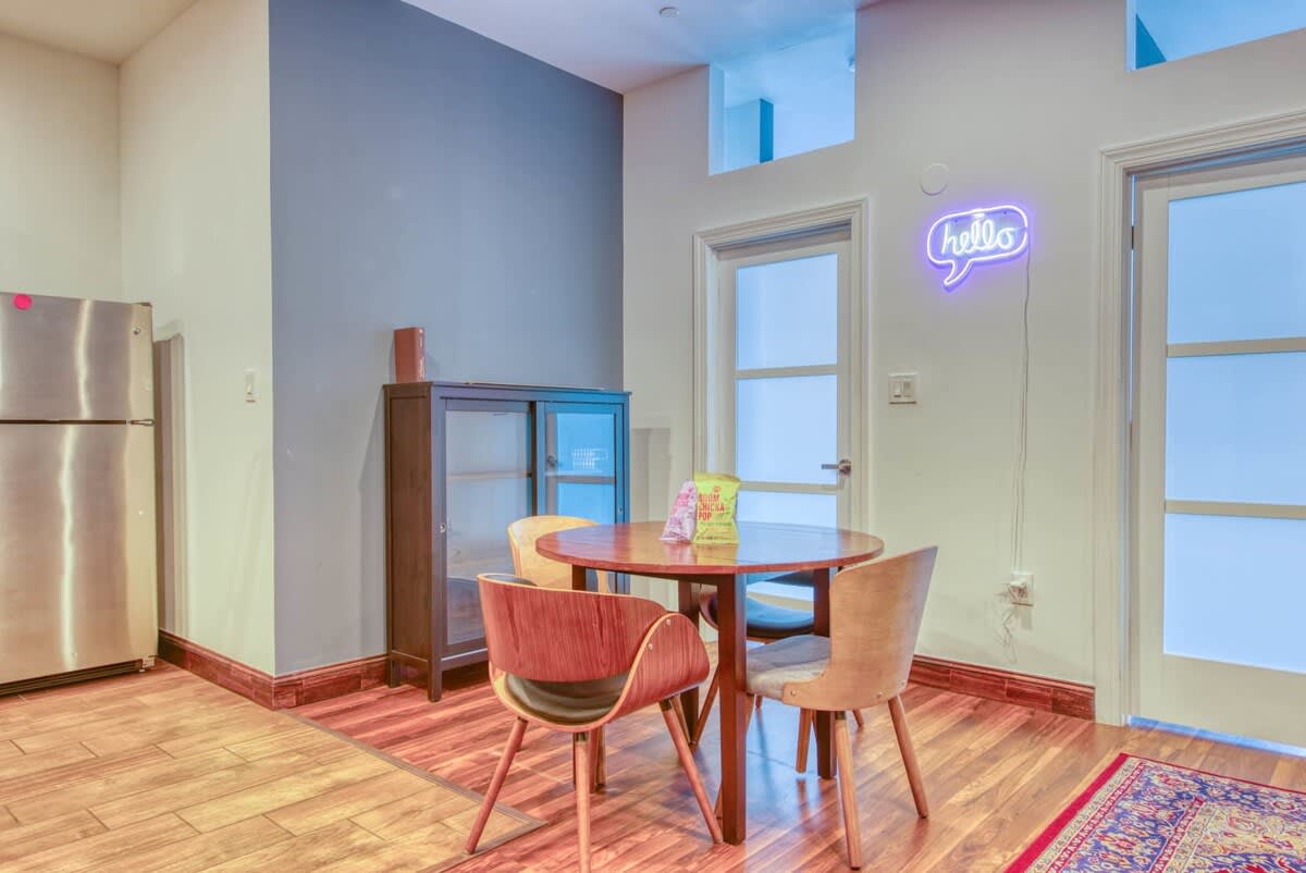A bright dining area with a round wooden table and four chairs, illuminated by a "hello" neon sign, adjacent to a dark display cabinet and a modern stainless steel refrigerator in the open-plan space.