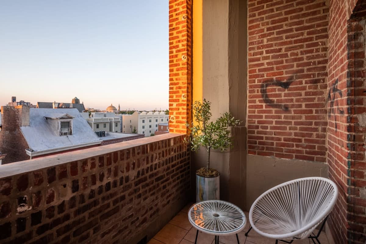 Small outdoor balcony with exposed brick walls, featuring a white woven chair and a small round table, overlooking city rooftops under a warm sunset glow. A potted plant adds a touch of greenery.