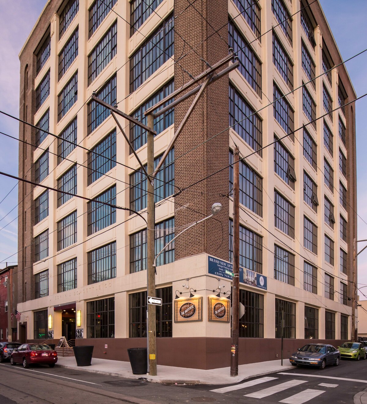 A tall, multi-story building with a facade of light-colored panels and brick, featuring numerous large, dark-framed windows, situated on an urban street corner with power lines and street signs.