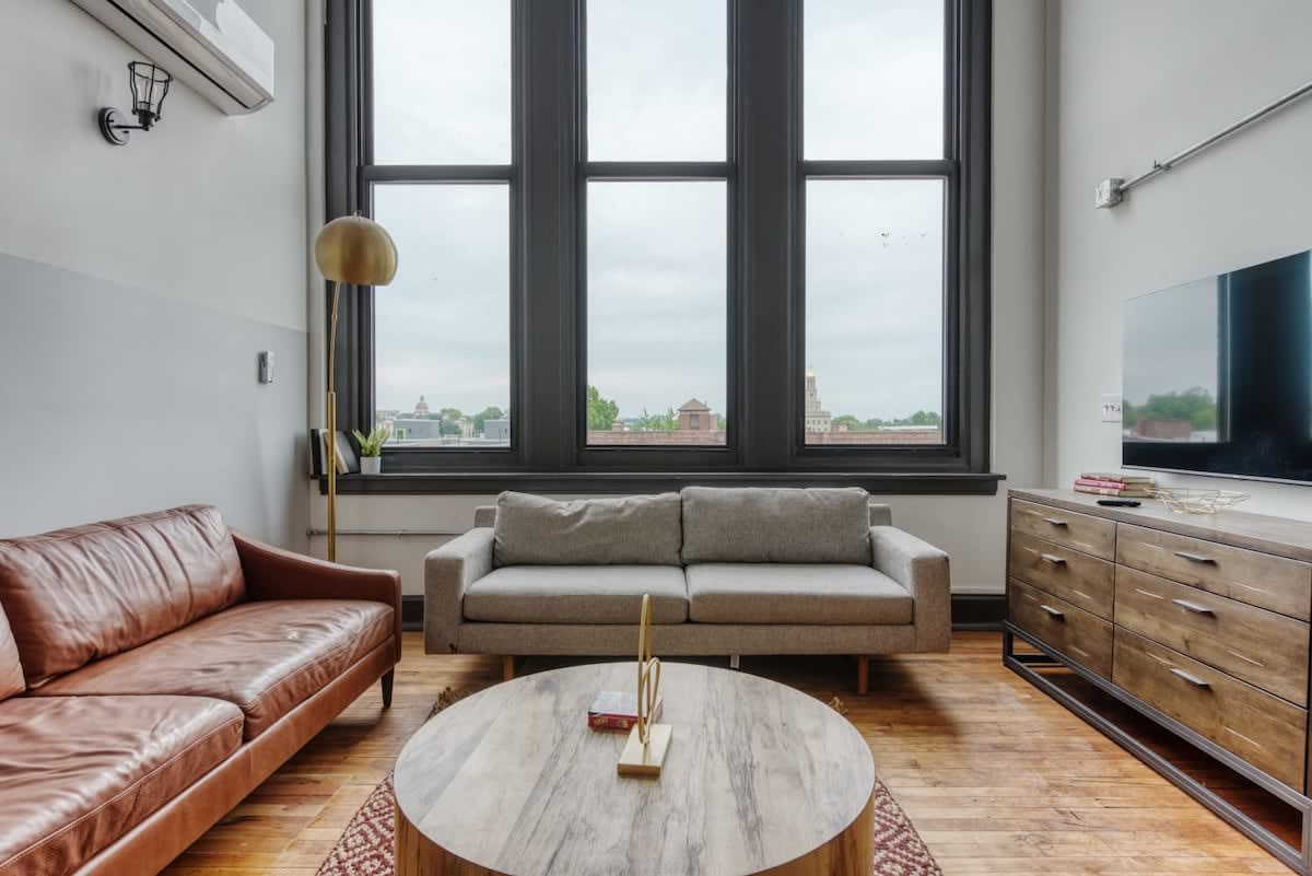 Bright living room with a brown leather sofa and a grey fabric sofa facing a round wooden coffee table, illuminated by natural light from tall, dark-framed windows.