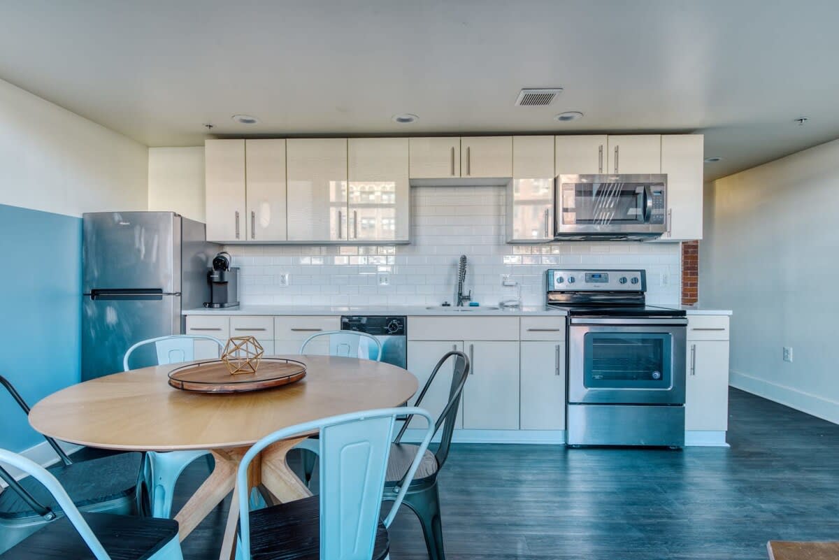 Bright, modern kitchen featuring white subway tile backsplash, light-colored flat-panel cabinets, and stainless steel appliances including a refrigerator, microwave, and oven. A round wooden dining table with light blue and black chairs is in the foreground.