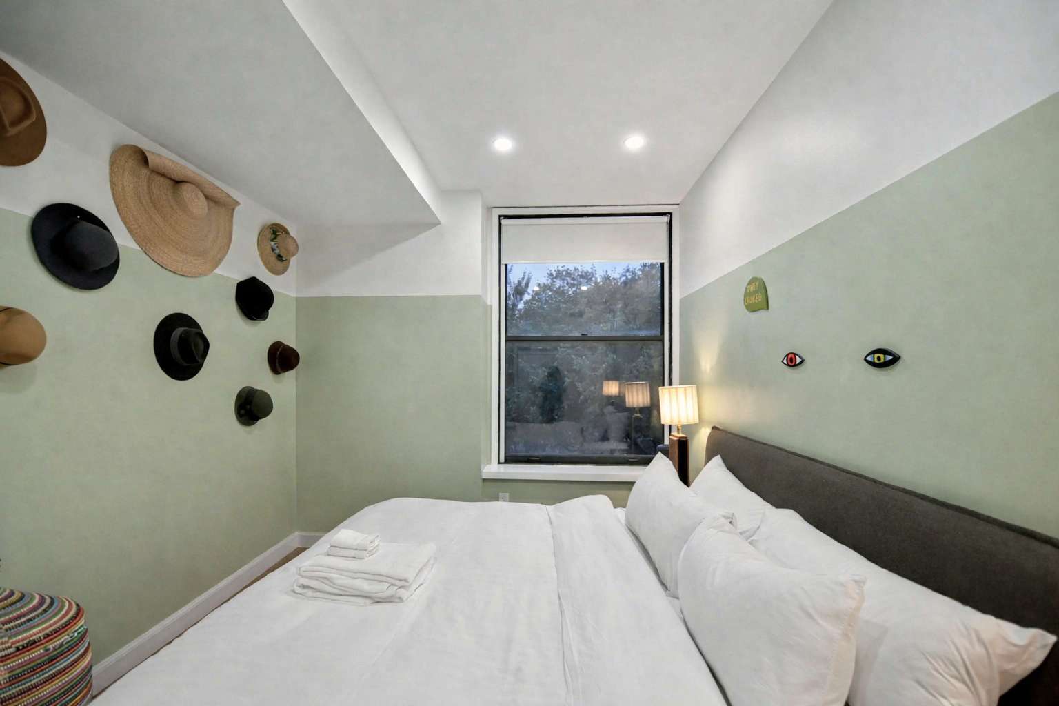 A bedroom with a queen-sized bed dressed in white linens is framed by a window, with walls painted in a two-tone light green and white scheme. Hats are decoratively hung on the green wall, adding a unique touch.