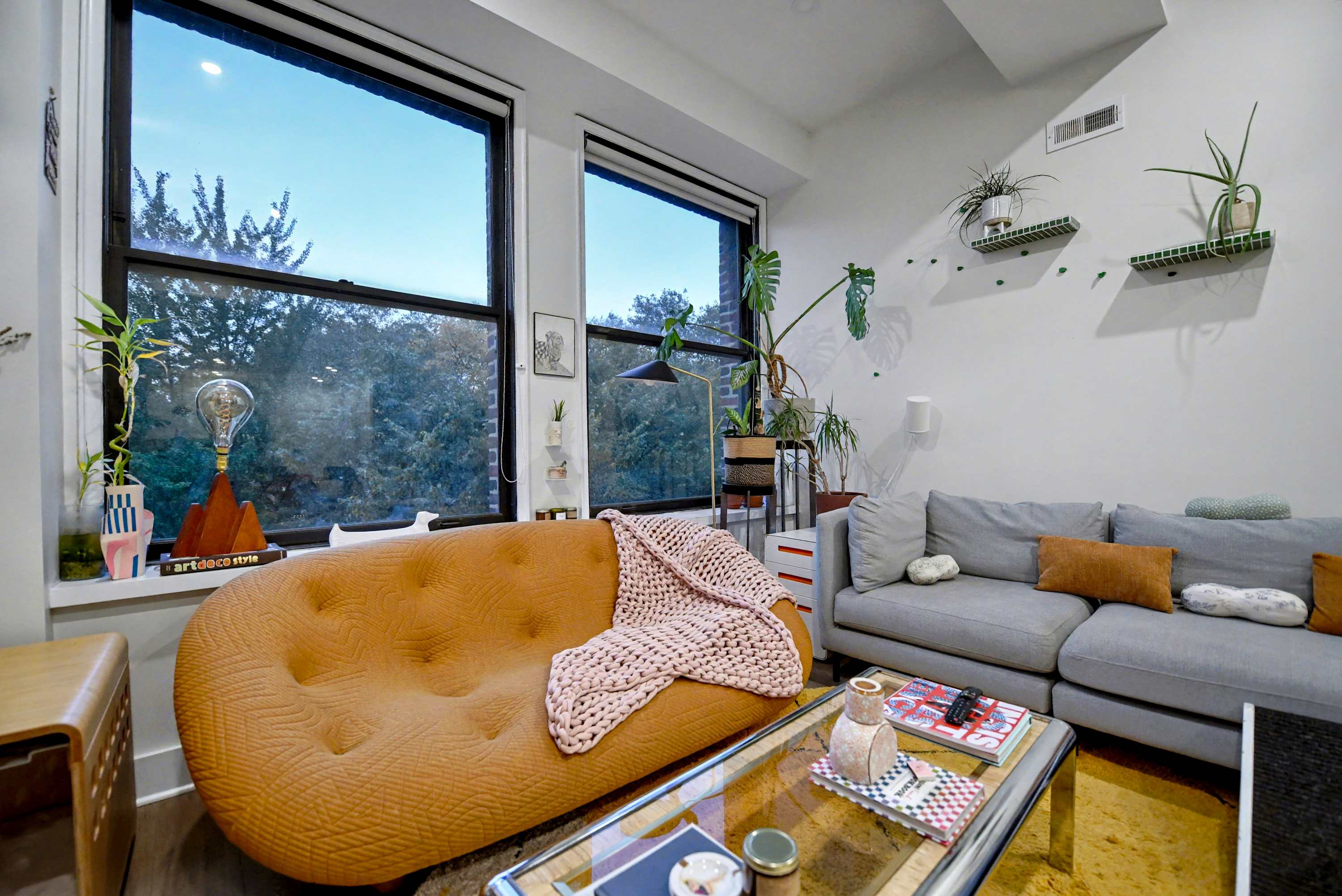 A cozy living room showcases an orange tufted sofa draped with a pink blanket, a gray sectional, and large windows overlooking trees. Various potted plants and decorative items adorn the window sills and shelves.