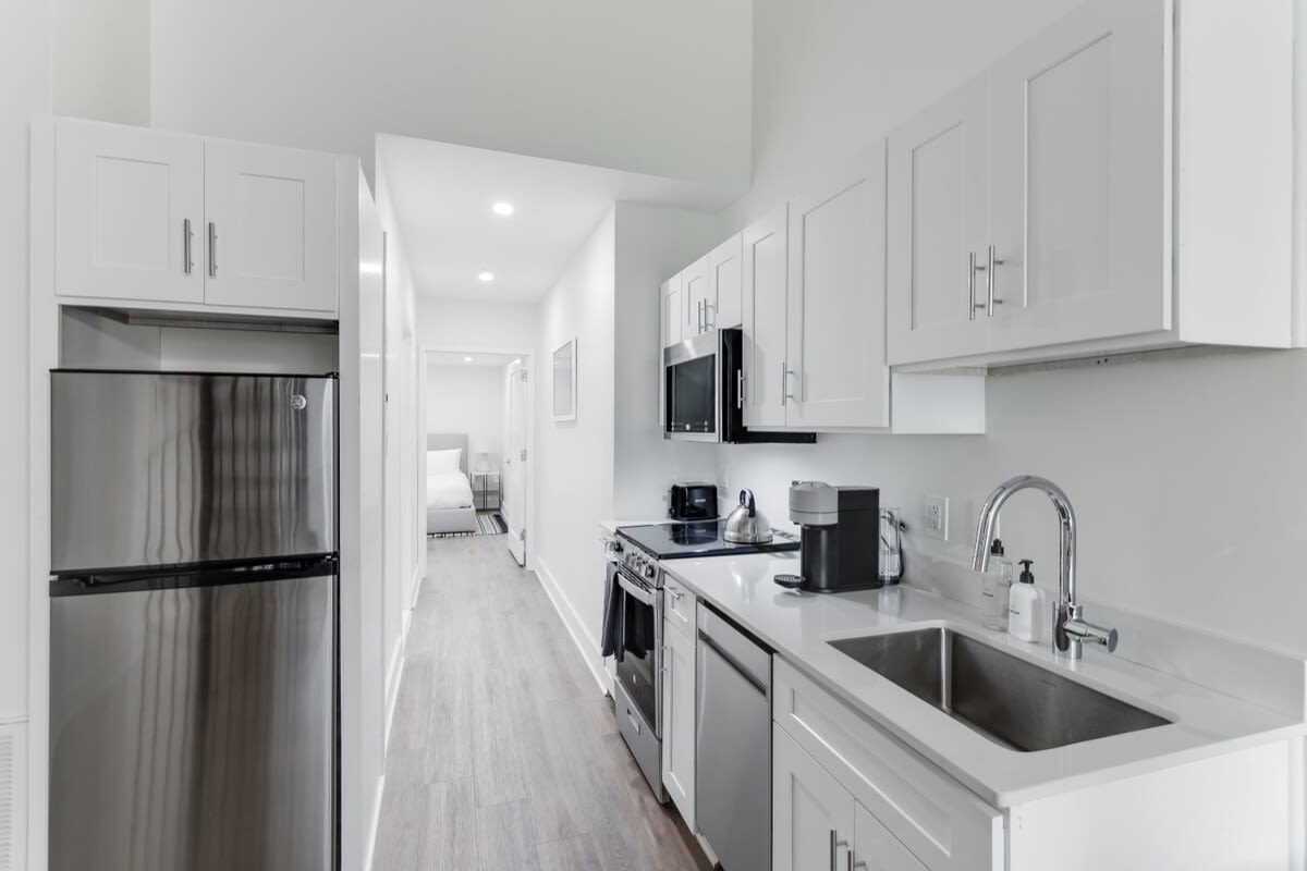 Bright, narrow kitchen with white shaker cabinets, white countertops, and stainless steel appliances including a refrigerator, microwave, and stove, with a hallway visible in the background.