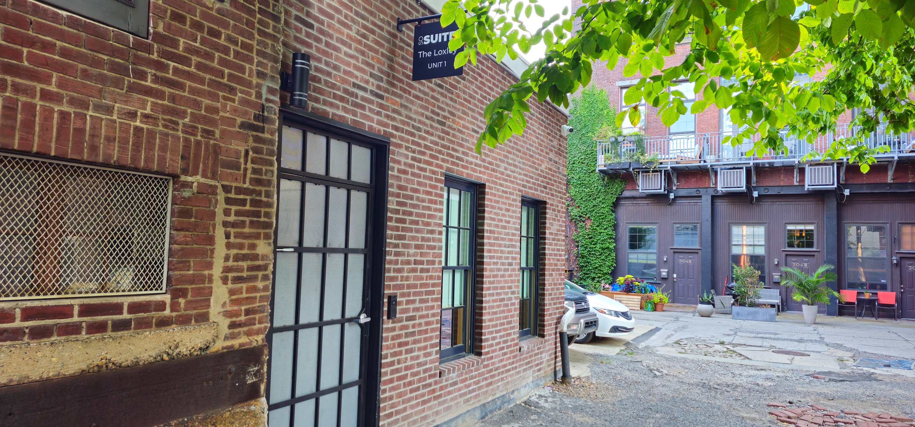 Exterior view of a brick building with black-framed windows and a black door, featuring a 'Sosuite' sign. Lush green foliage hangs overhead, and a paved courtyard is visible in front.