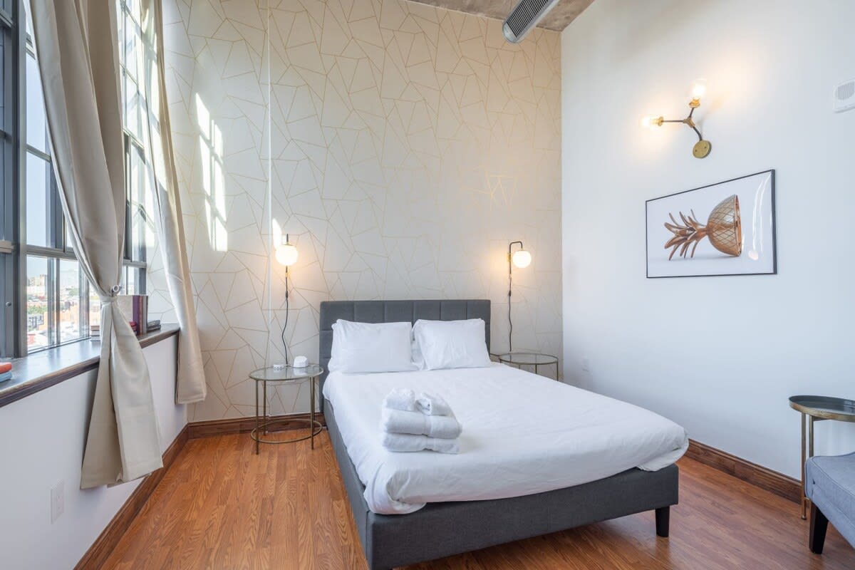 Cozy bedroom with a gray upholstered bed, white bedding, two glass-top nightstands with modern lamps, and a subtle gold geometric patterned wallpaper on one wall, illuminated by large industrial-style windows with light curtains.
