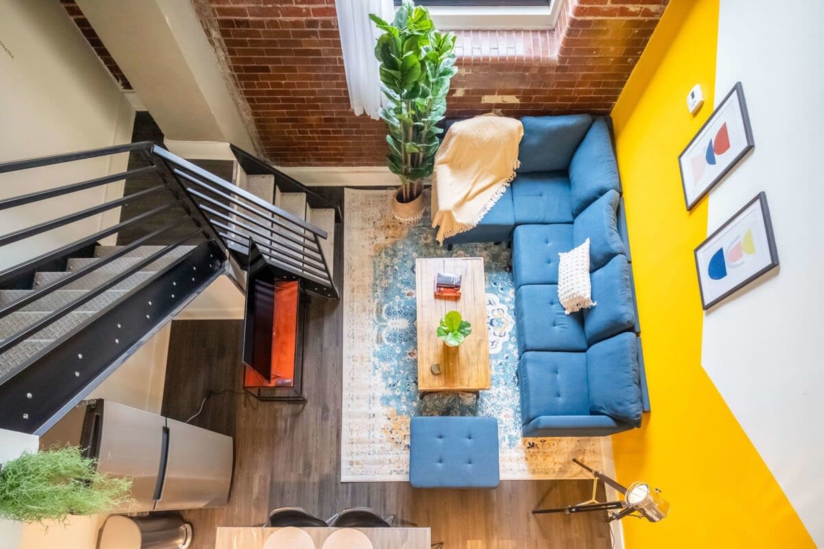 Overhead view of an open-concept loft space, showcasing a blue sectional sofa on a patterned rug, a wooden coffee table, and a dining area with a table and black chairs. The kitchen is visible in the background, and a black metal staircase leads to an upper level.