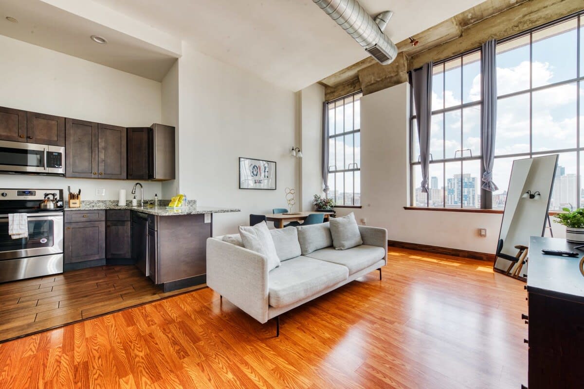 An open-plan kitchen and living area, showcasing a contemporary kitchen with dark wood cabinetry, speckled granite countertops, and stainless steel appliances. The space flows into a living area with a gray sofa and large windows.