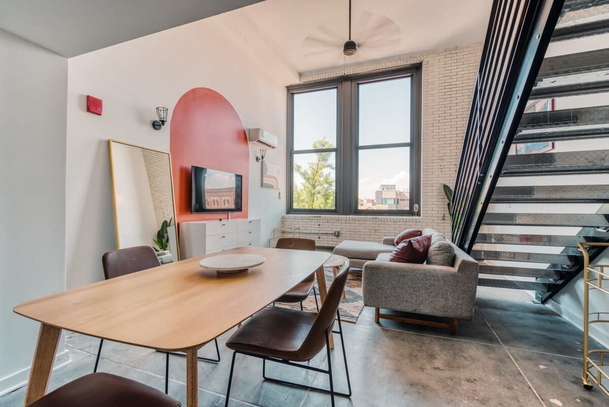 Inviting dining area featuring a round wooden table with four light wood chairs with woven seats, set on a polished concrete floor. In the background, the living room offers a gray sofa, a white media console with a TV, and large windows, all under a ceiling fan.