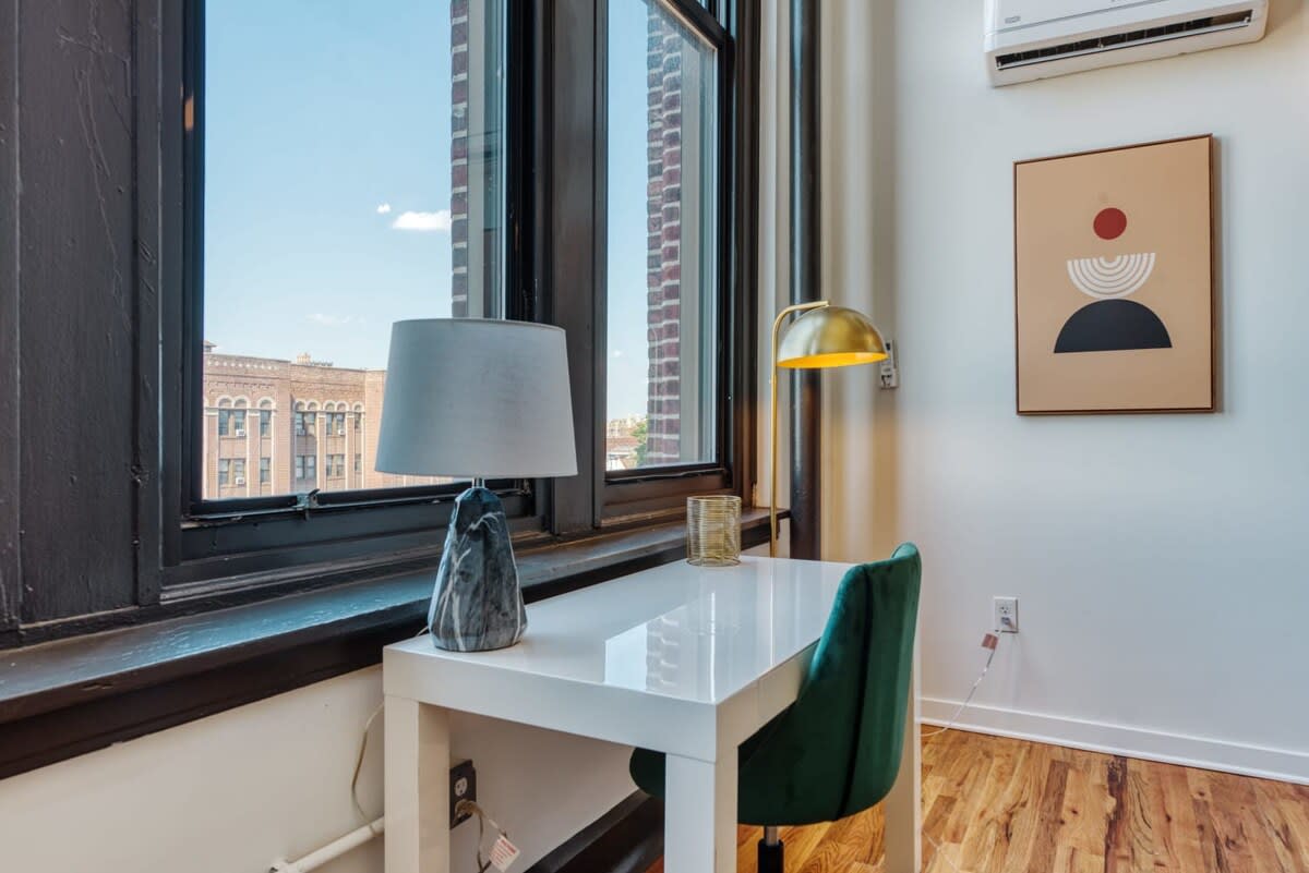 Dedicated workspace featuring a white desk and a green velvet office chair, positioned by large windows offering city views. A modern table lamp and a gold floor lamp illuminate the area.