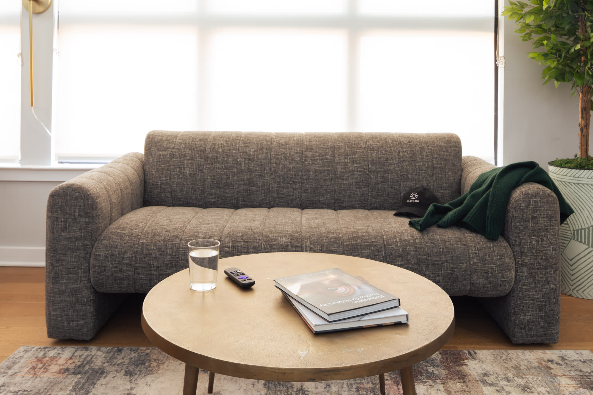 A close-up of a gray textured sofa with a green throw blanket and a cap, a round gold coffee table with books, a remote, and a glass of water, set on a patterned area rug. Large windows with white roller shades are in the background.