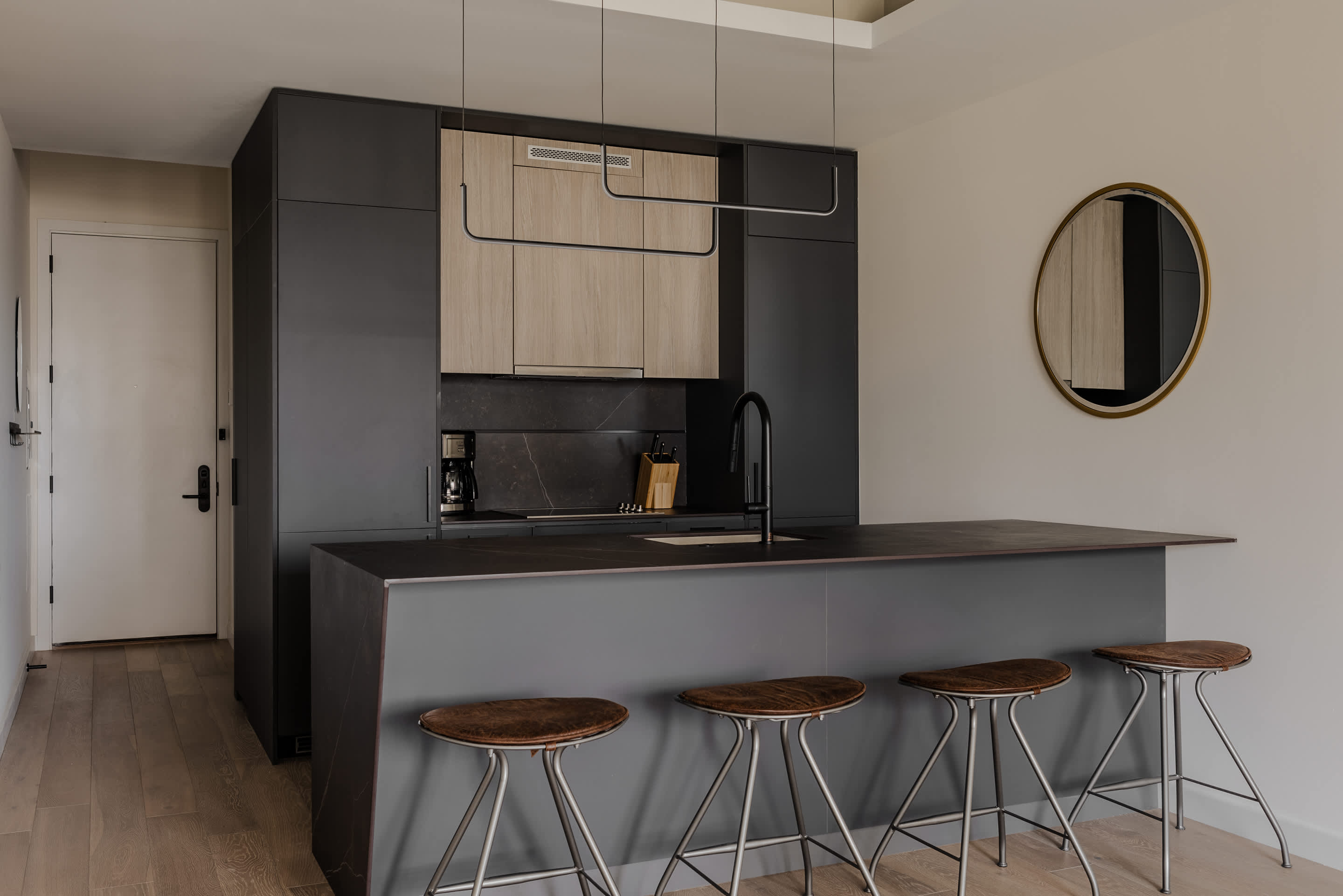 Sleek modern kitchen featuring dark grey flat-panel cabinetry, light wood upper cabinets, a large island with a dark countertop and four bar stools, illuminated by contemporary linear pendant lights.
