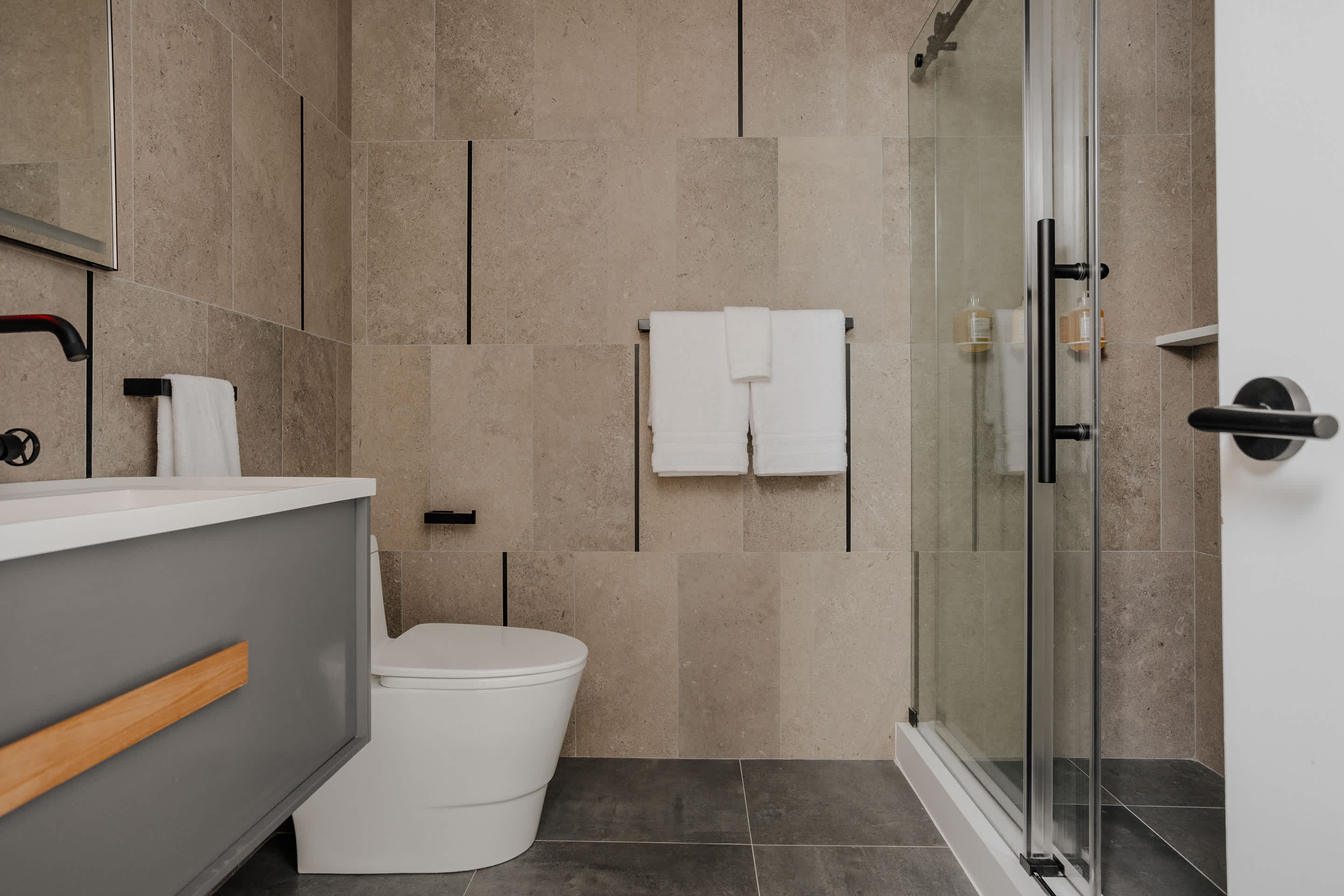 Modern bathroom with light beige rectangular tiled walls, a grey floating vanity with a white sink, a white toilet, and a glass-enclosed shower, accented with black fixtures.