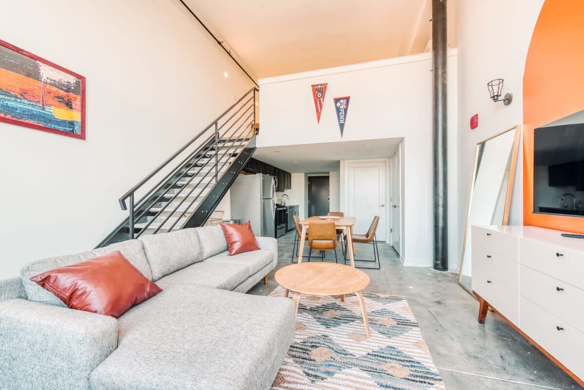 Bright and spacious loft living room with a light gray L-shaped sectional sofa, red accent pillows, and a round wooden coffee table. Beyond the living area, a dining table with four chairs and a compact kitchen are visible, all set against white walls and concrete floors.