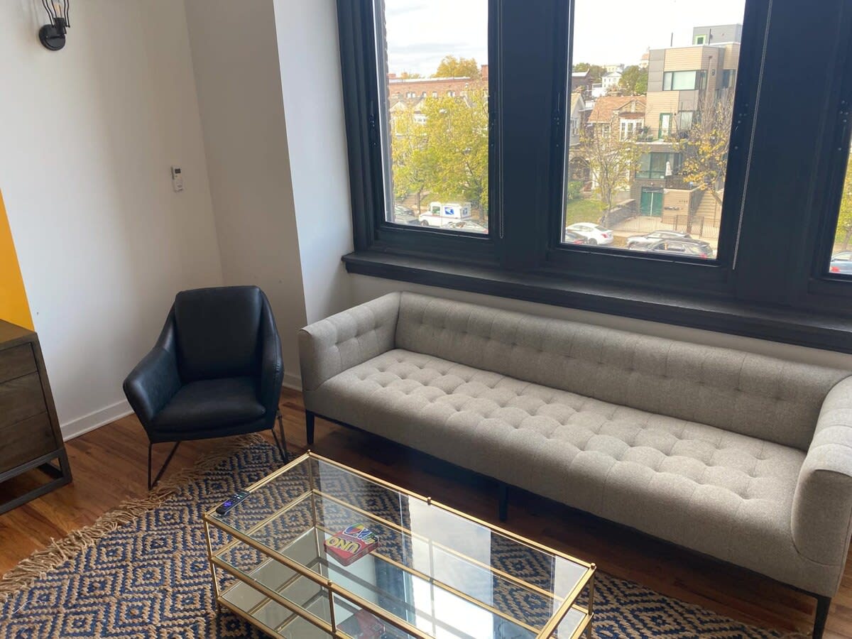 Close-up of the living area with a light gray tufted sofa, a black armchair, and a gold-framed glass coffee table on a patterned rug, positioned in front of large black-framed windows overlooking the city.