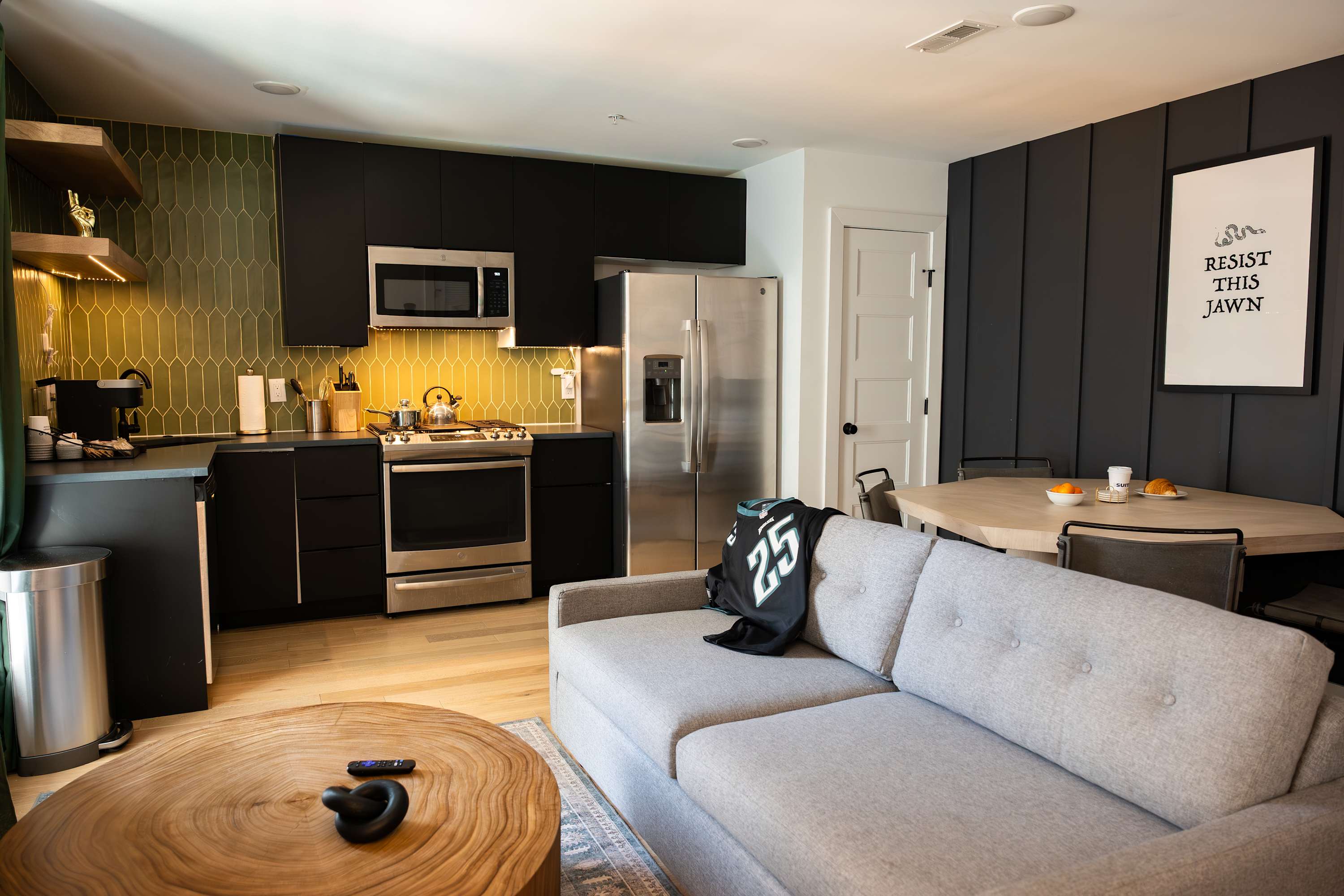 A stylish kitchen equipped with black cabinetry, stainless steel appliances, and a distinctive green geometric tile backsplash, adjacent to a dining area and a gray sofa.