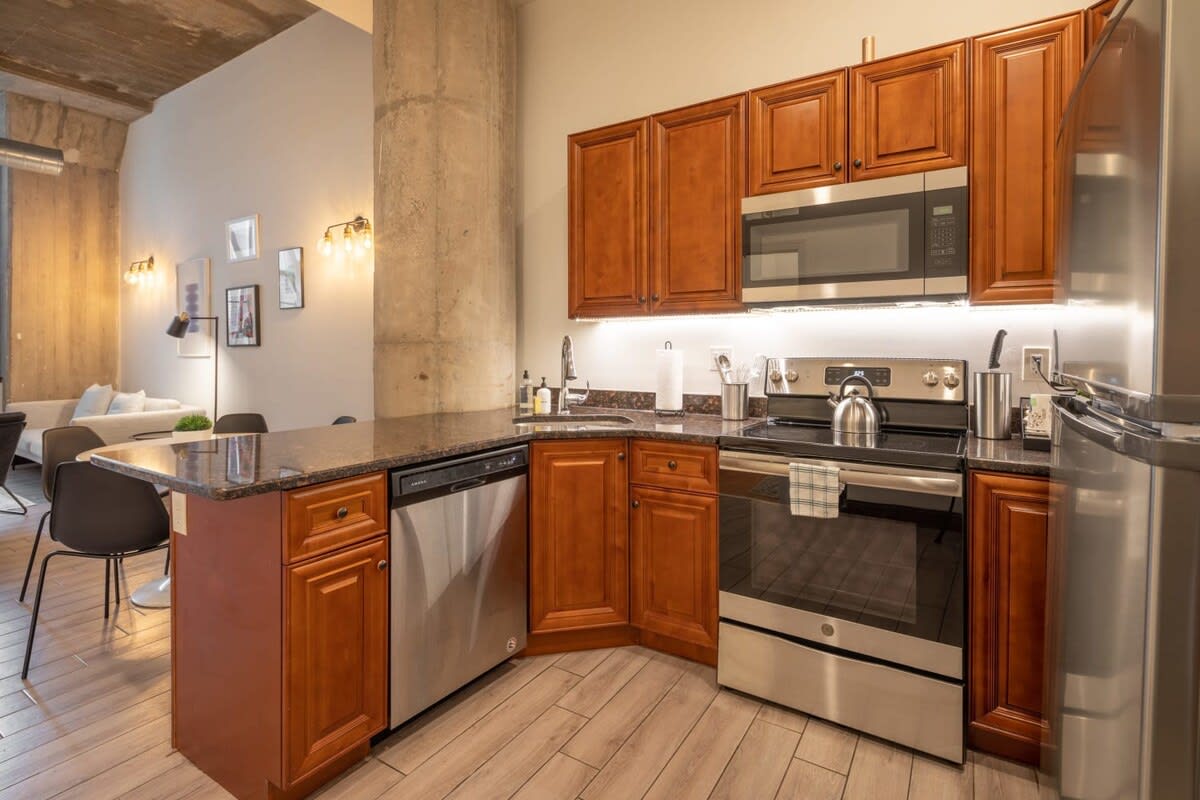 Fully equipped kitchen with warm wooden cabinetry, dark granite countertops, and stainless steel appliances including a microwave, oven, dishwasher, and refrigerator, with under-cabinet lighting.