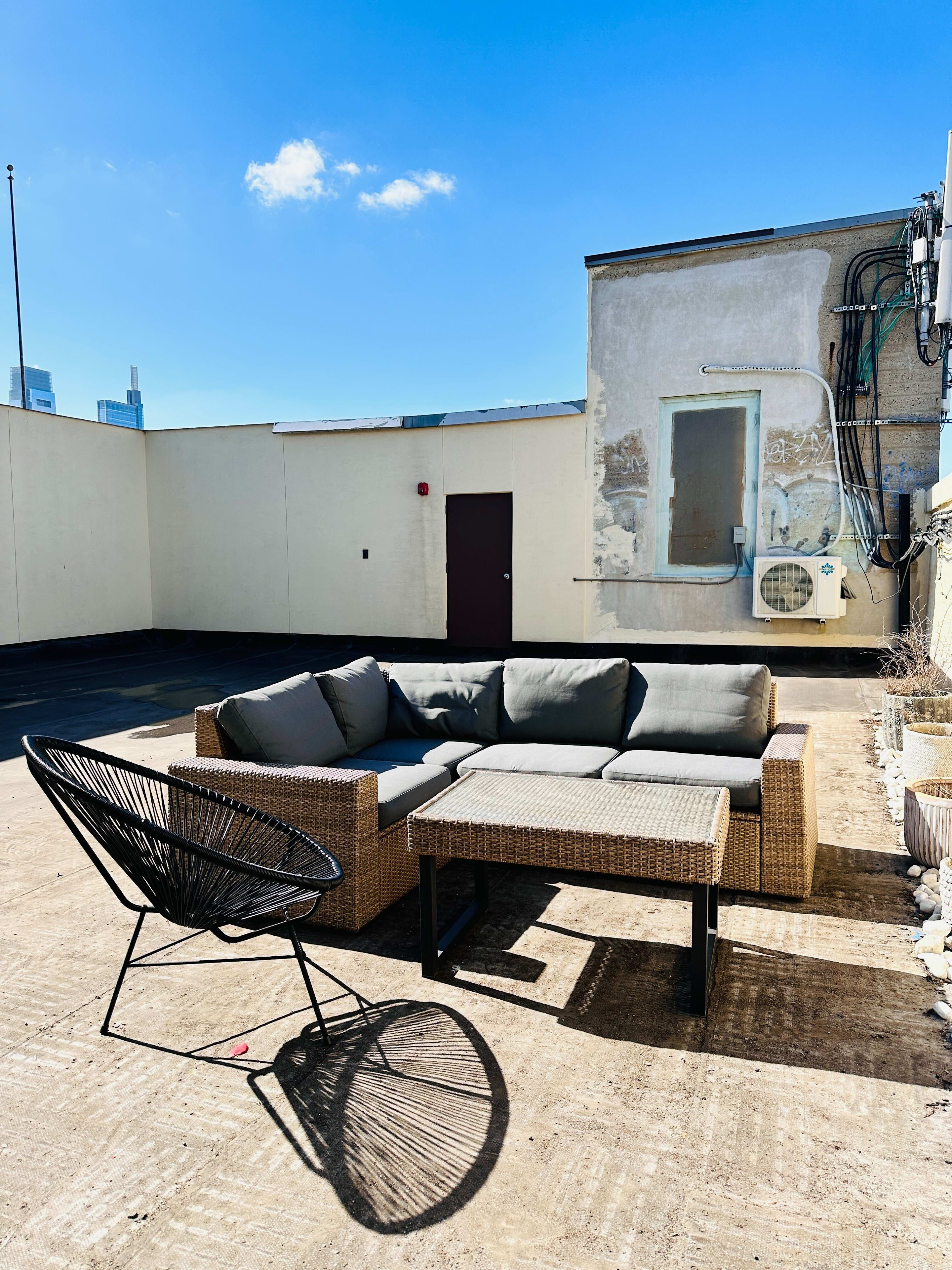 Sunny rooftop patio with a wicker sectional sofa and a matching coffee table, along with a black string chair. The concrete floor is visible, and in the background, there's a building wall with an air conditioning unit and a distant city skyline.