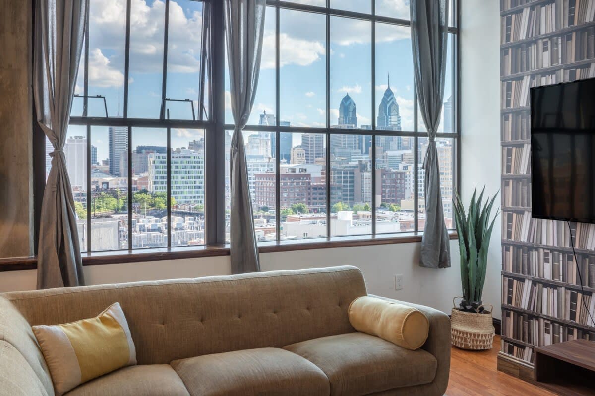 Close-up view from the living room showing large industrial-style windows with grey curtains, offering a panoramic city skyline view with prominent skyscrapers.
